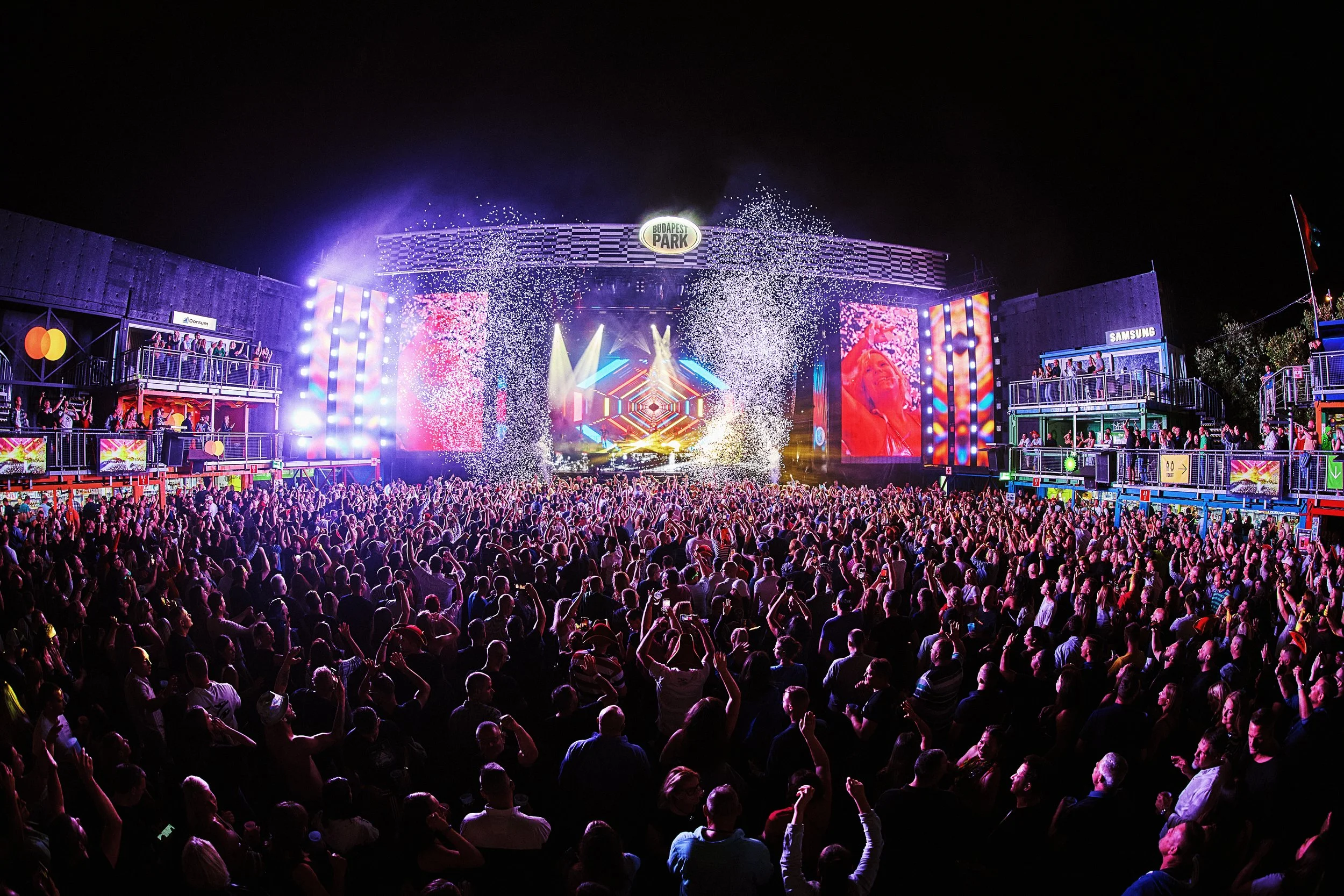 Large outdoor concert at night with a stage, bright lights, video screens, and a crowd of people celebrating with confetti.