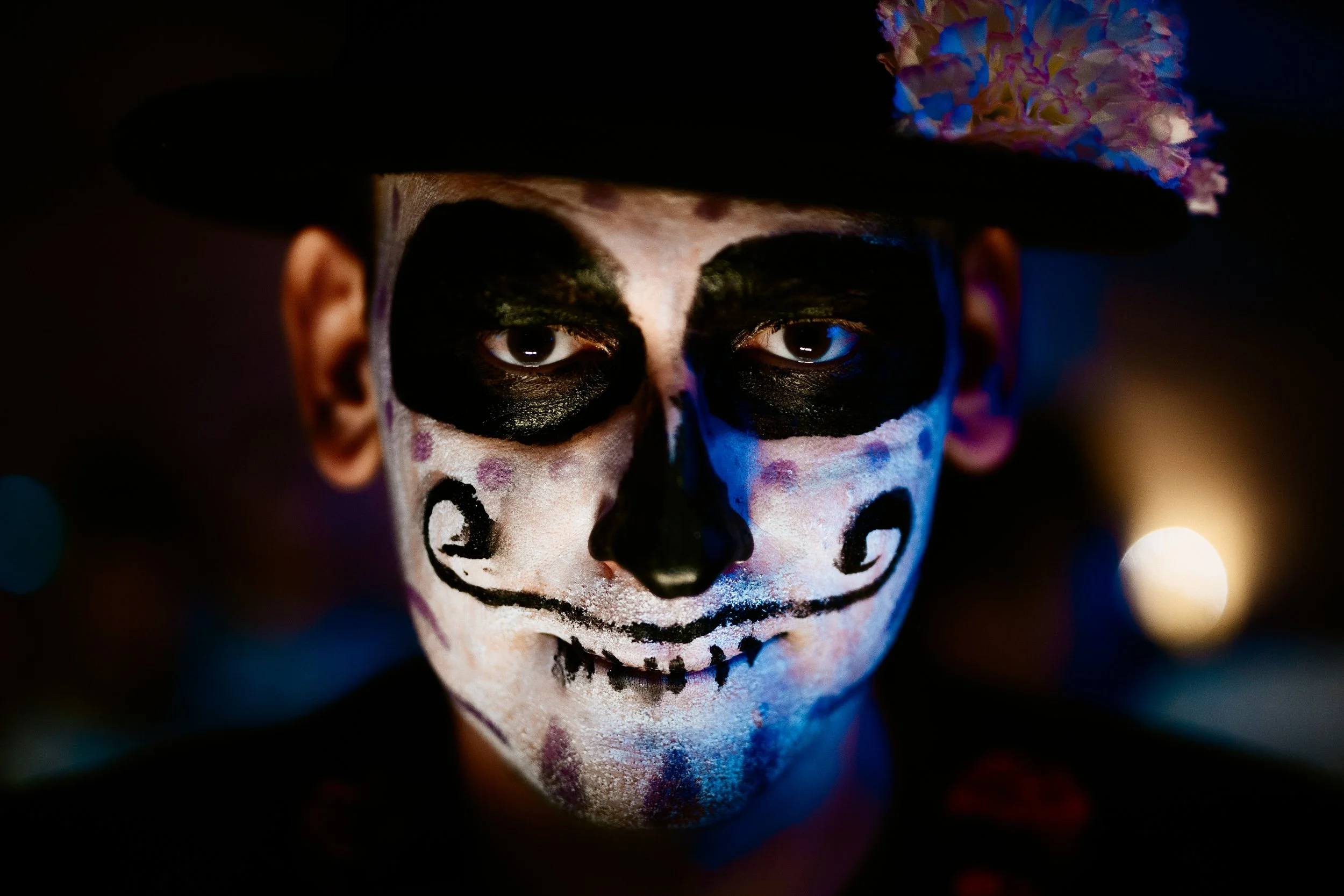 Person with painted face resembling a skeleton, wearing a black hat with colorful decorations, in dim lighting.