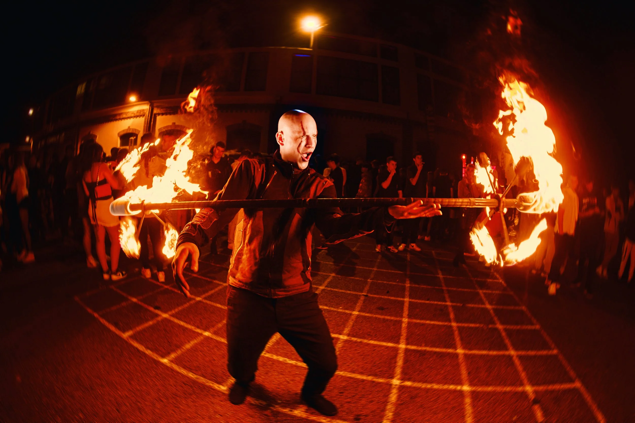 A man performing fire juggling at night during a festival, with a crowd of onlookers behind him.
