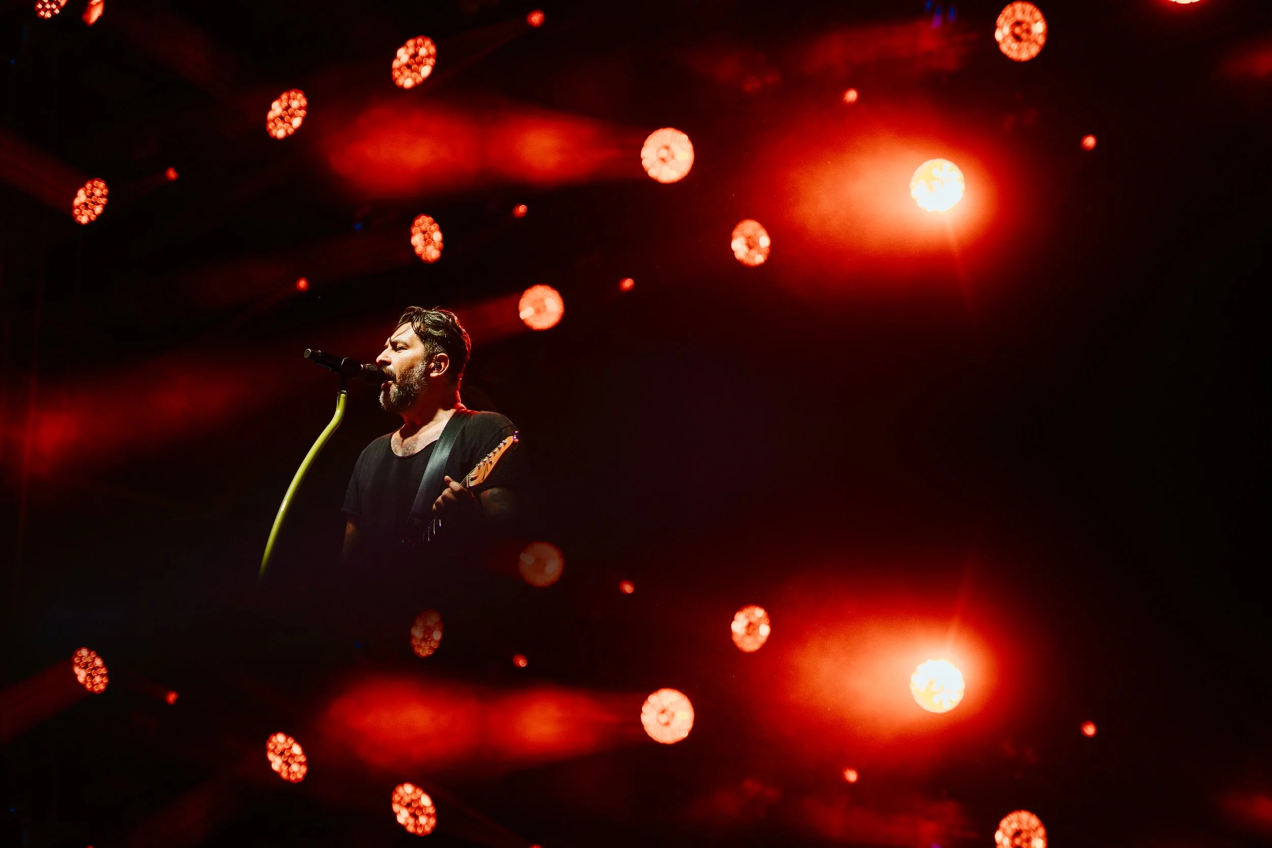 Musician singing into a microphone and playing guitar on stage with red stage lights.