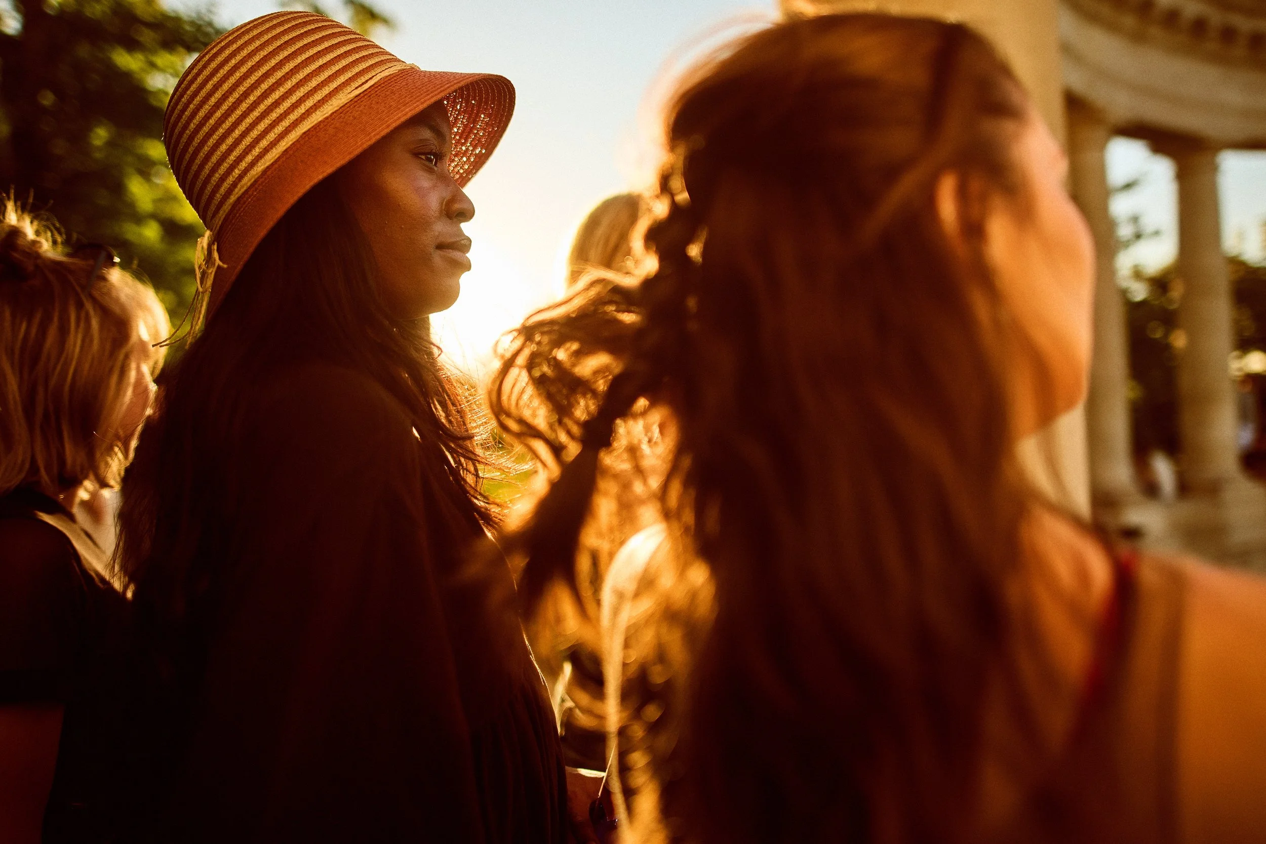 A group of women standing outdoors during sunset, with one woman wearing a large striped sun hat and looking contemplative.