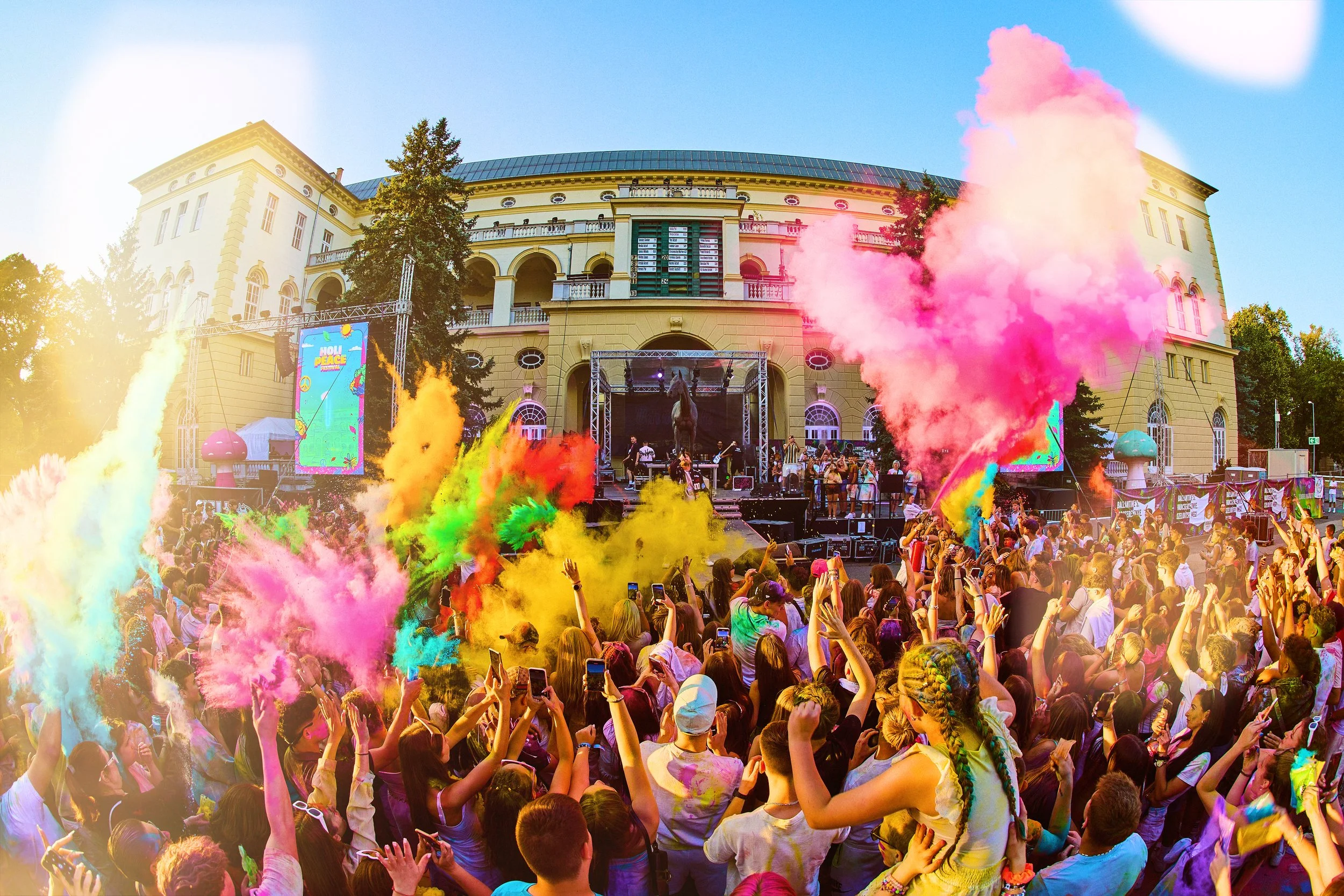 Crowd of people enjoying a colorful outdoor music festival with performers on stage, colorful powder clouds in the air, and a historic building in the background.