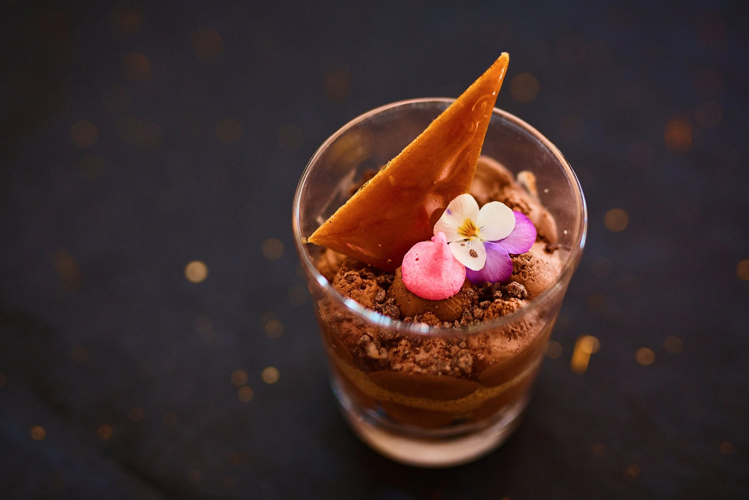 Chocolate mousse dessert topped with a cookie, edible flowers, and pink meringue in a glass