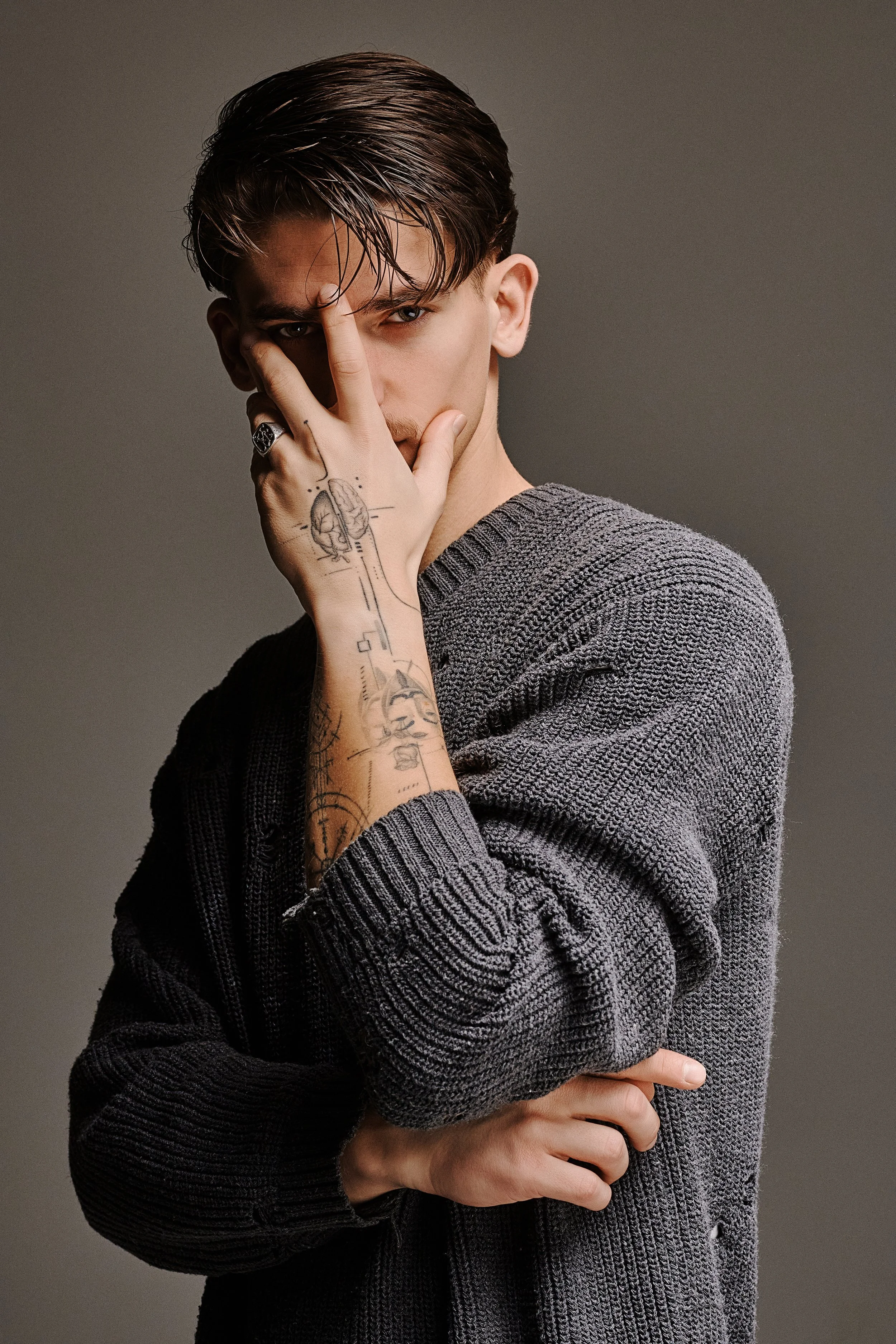 A young man with slicked-back brown hair and tattoos on his left hand and forearm, wearing a gray knitted sweater, is posing with his hand covering part of his face while looking directly at the camera against a plain gray background.