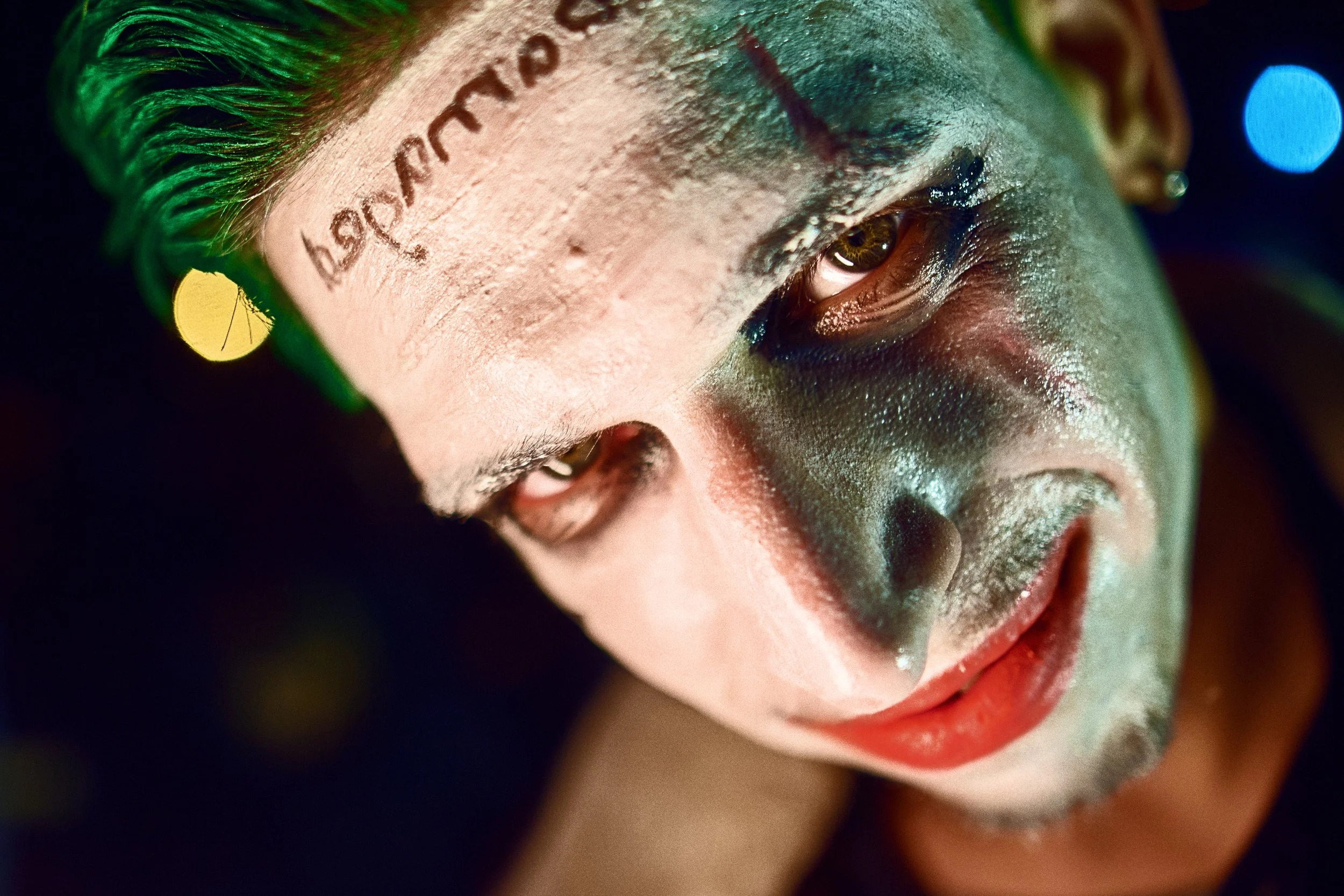 Close-up of a person with Joker-style makeup including white face paint, red lipstick, green hair, and a tattoo on the forehead, with colorful lights in the background.
