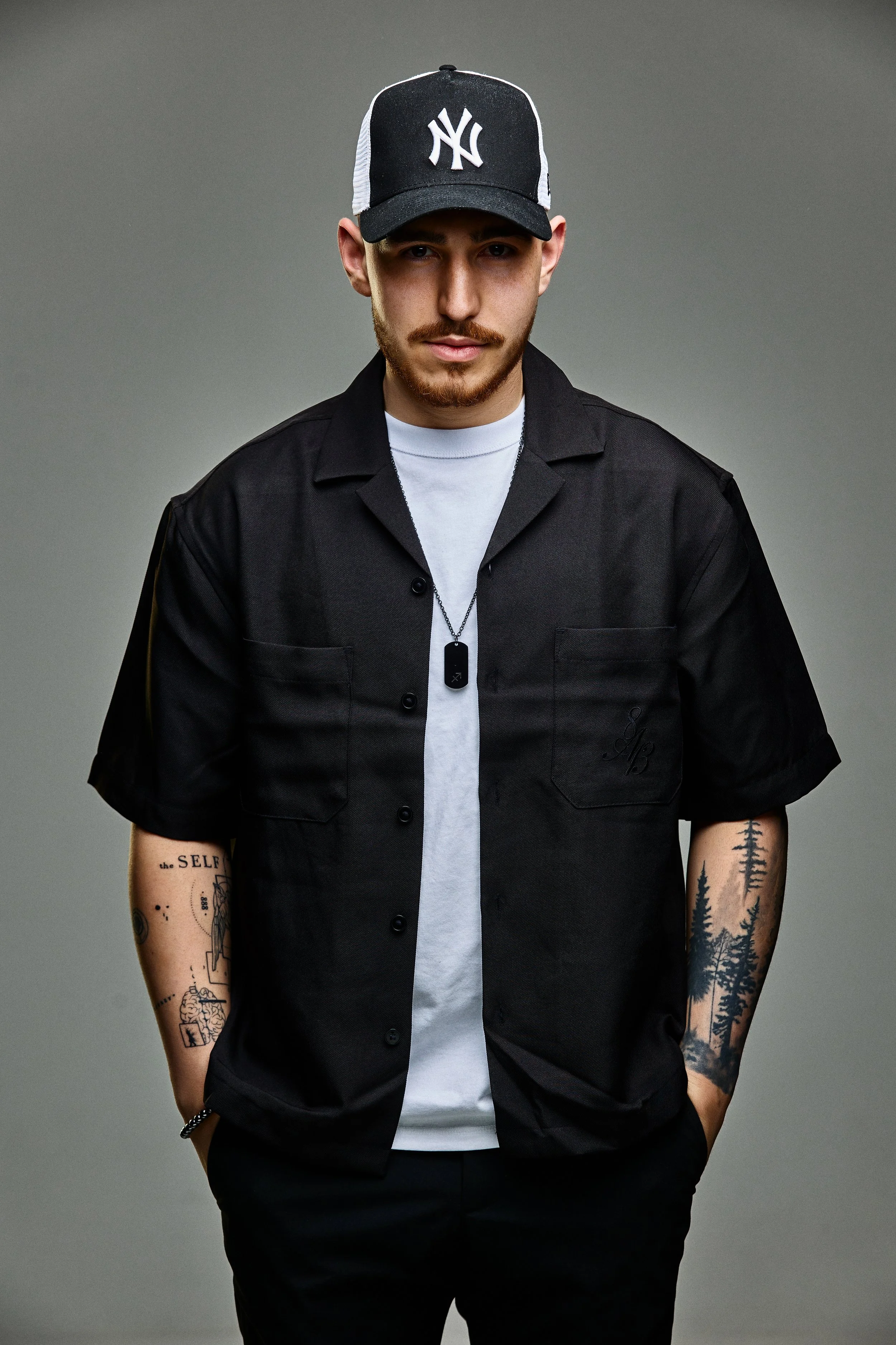Portrait of a young man with a mustache and beard, wearing a black New York Yankees baseball cap, black short-sleeve button-up shirt with embroidered initials, white T-shirt underneath, dog tag necklace, and tattoos on both arms against a gray backgr