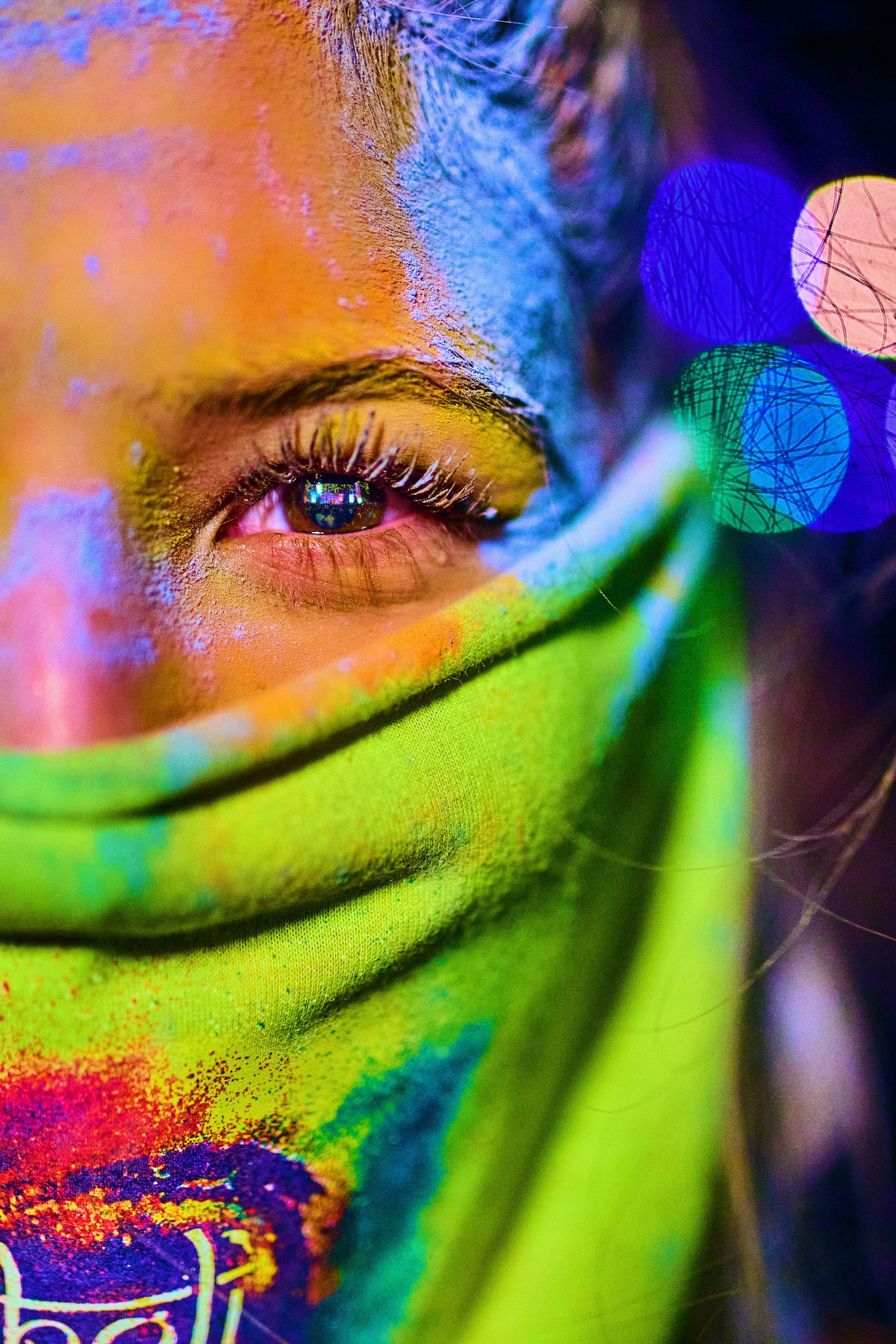 Close-up of a person's eye with colorful neon face paint in green, yellow, blue, and orange, partially covered by a green fabric, and vibrant bokeh lights in the background.