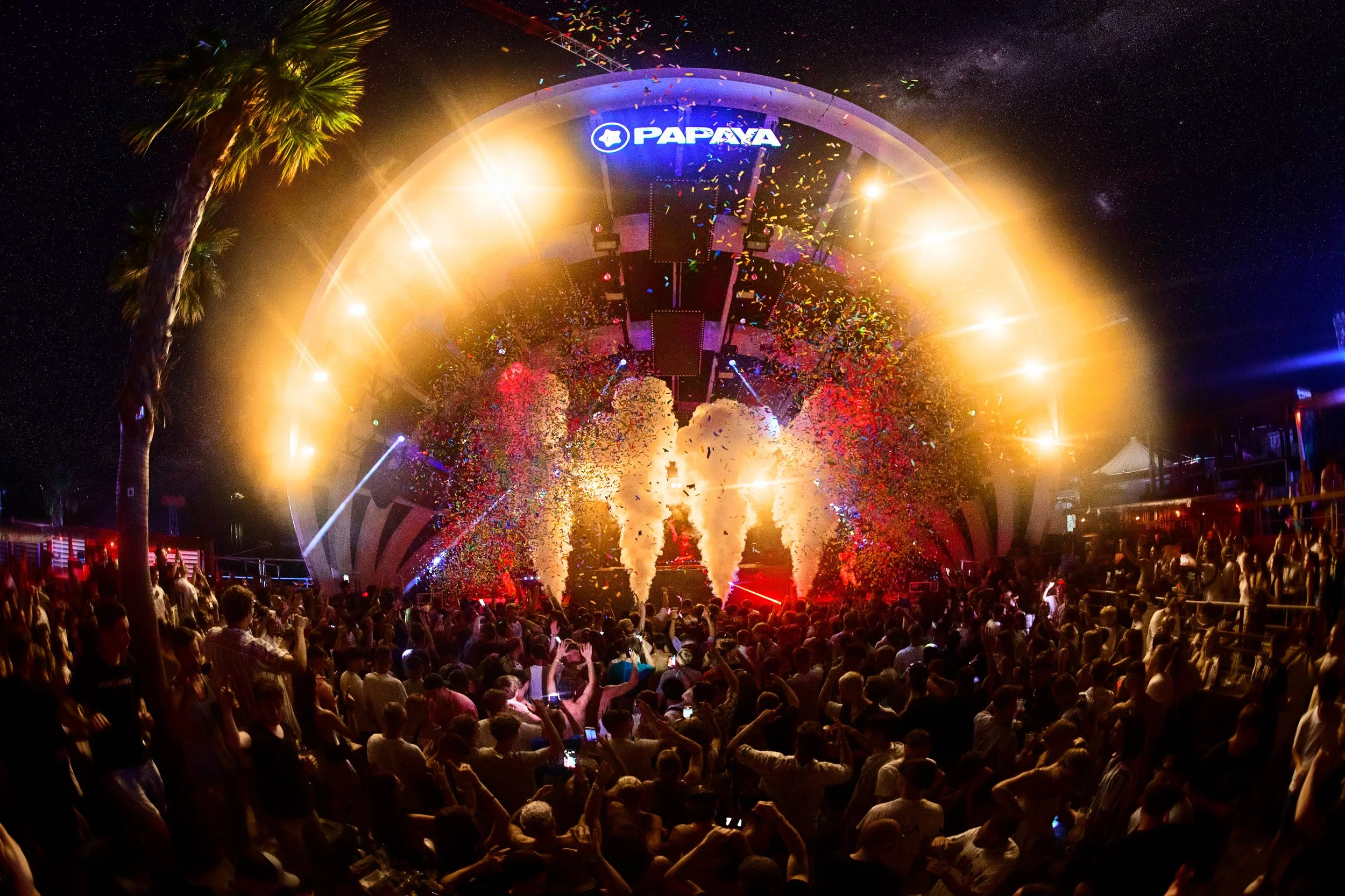 A nightclub or concert venue with a large crowd of people dancing and enjoying the show. The stage features bright lights, smoke, and confetti, with the brand name 'PAPAYA' illuminated at the top. The scene is energetic and vibrant with colorful ligh
