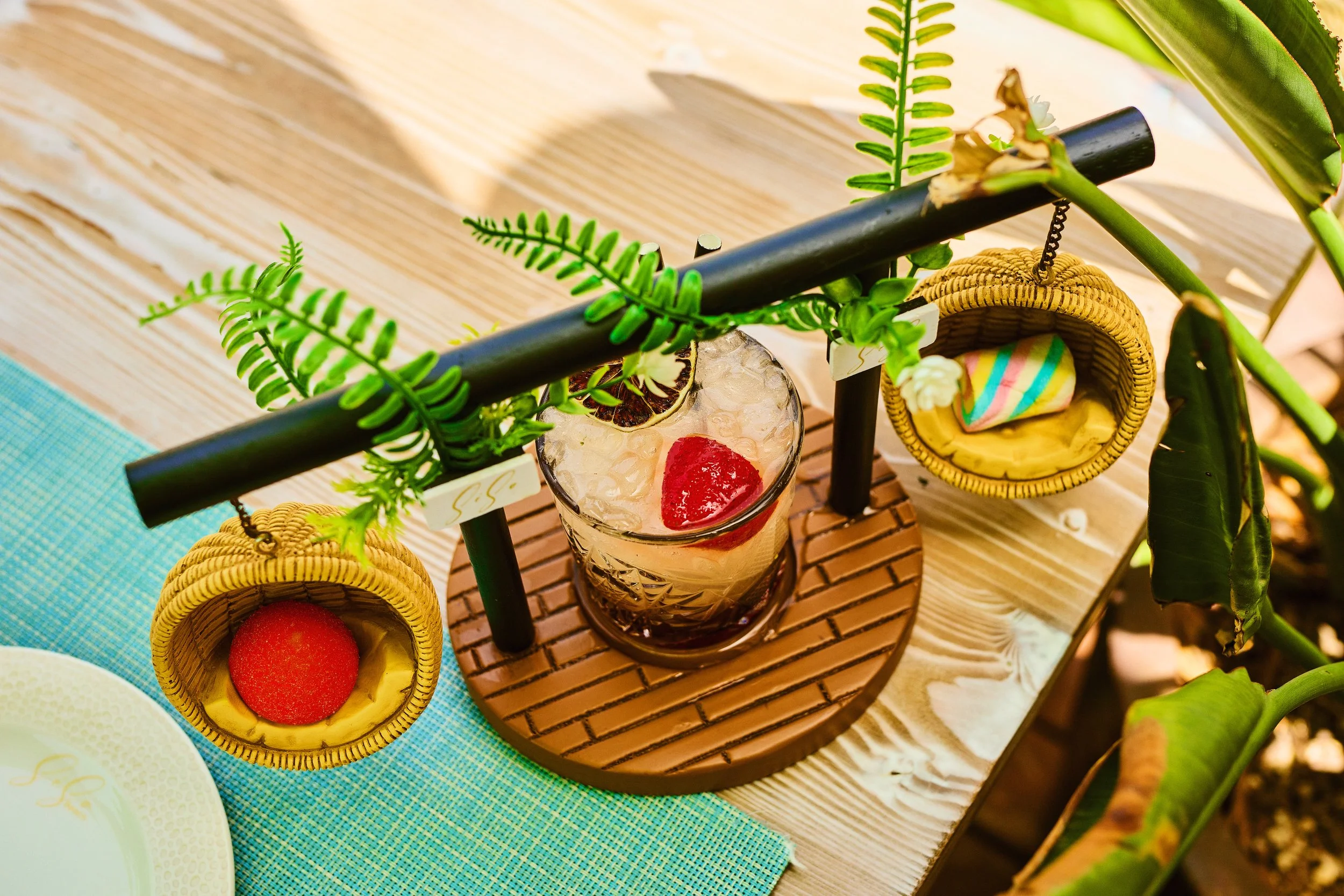 A colorful drink with ice and a strawberry inside on a wooden table, surrounded by small woven baskets with colorful candies, and decorated with green artificial fern leaves.
