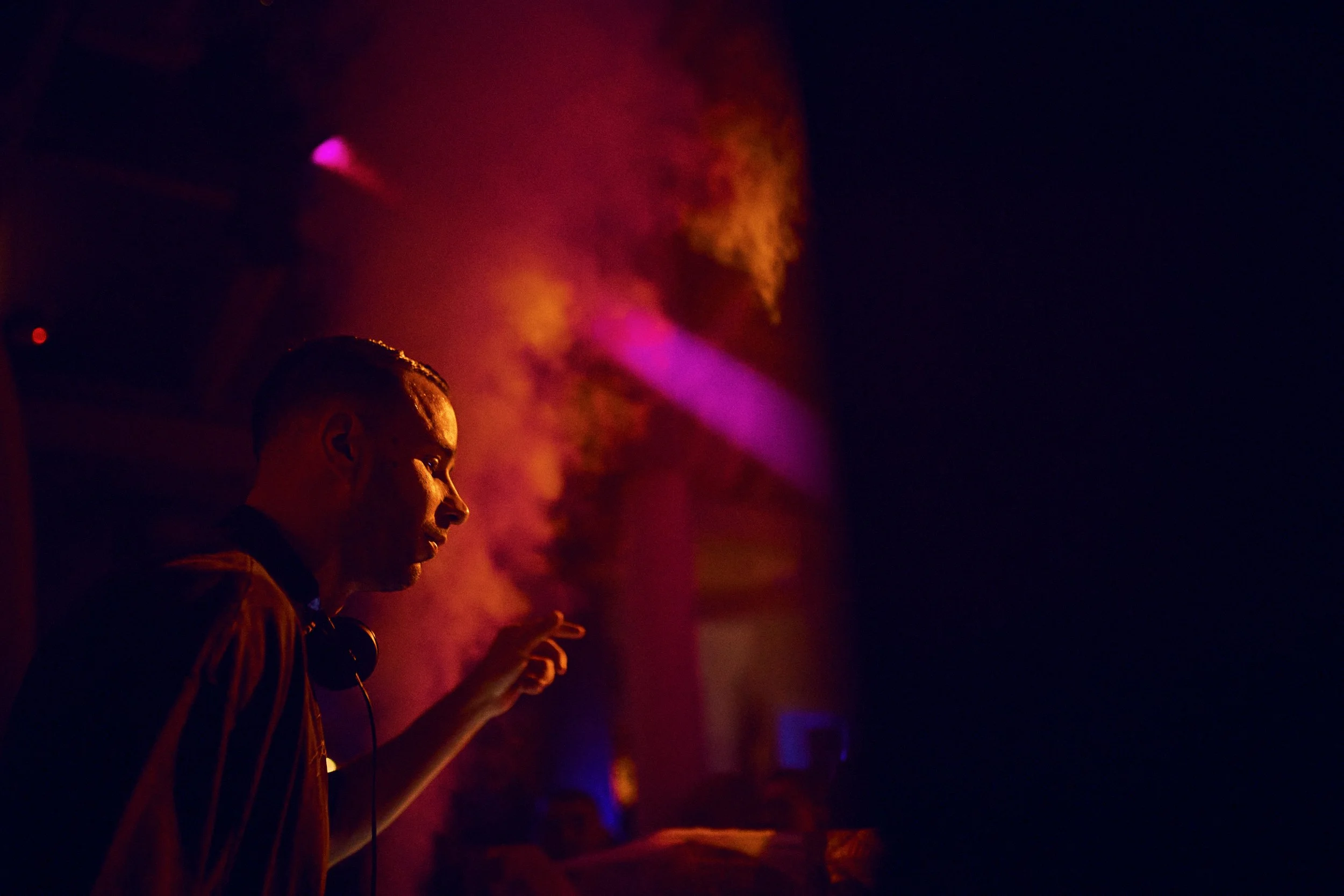 A DJ performing at a nightclub with vibrant purple and orange lighting, wearing headphones around his neck, and pointing with his right hand.