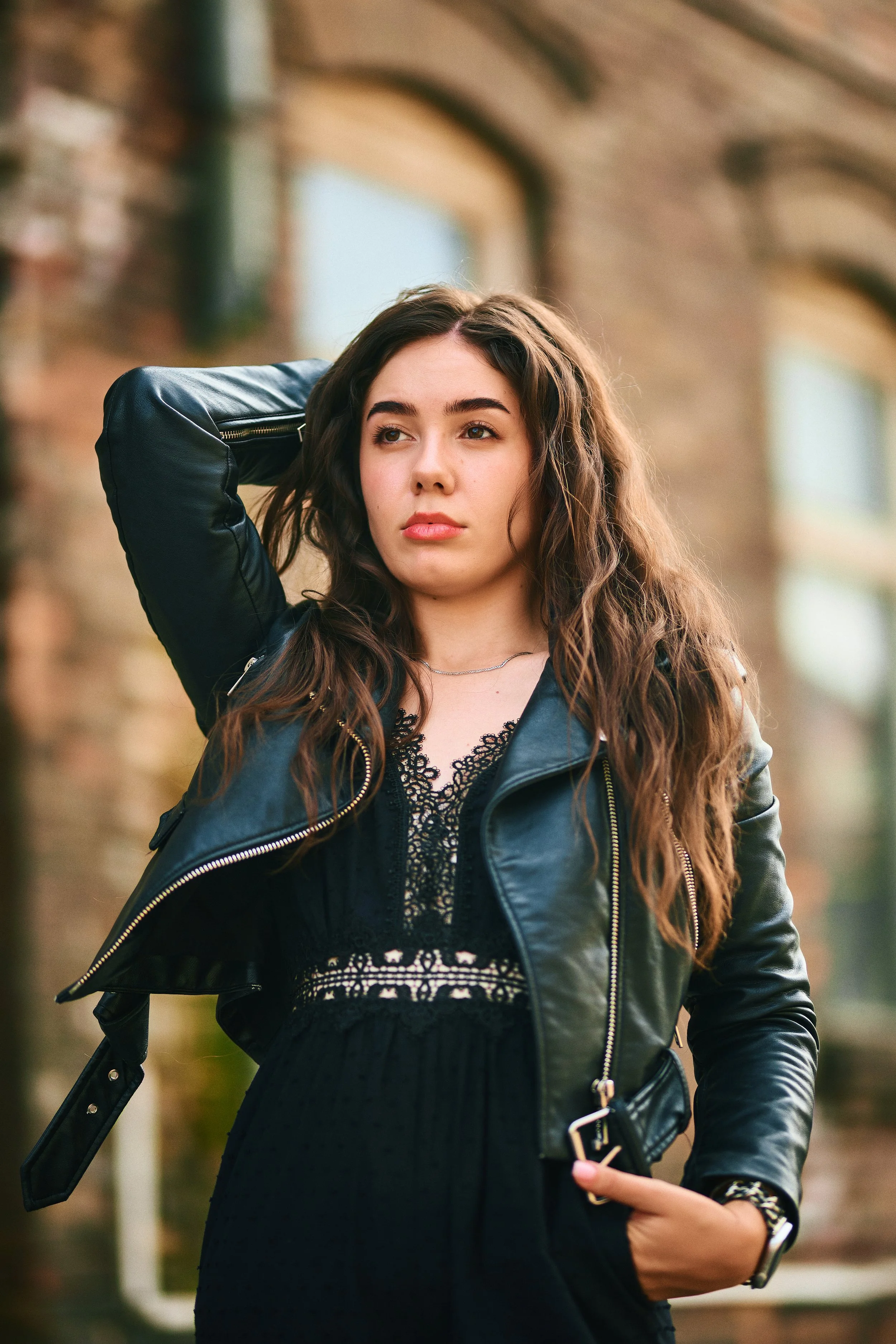 Young woman with long wavy brown hair wearing a black dress and black leather jacket, standing outdoors against a brick building, looking off to the side with one hand in her pocket and the other resting on her head.