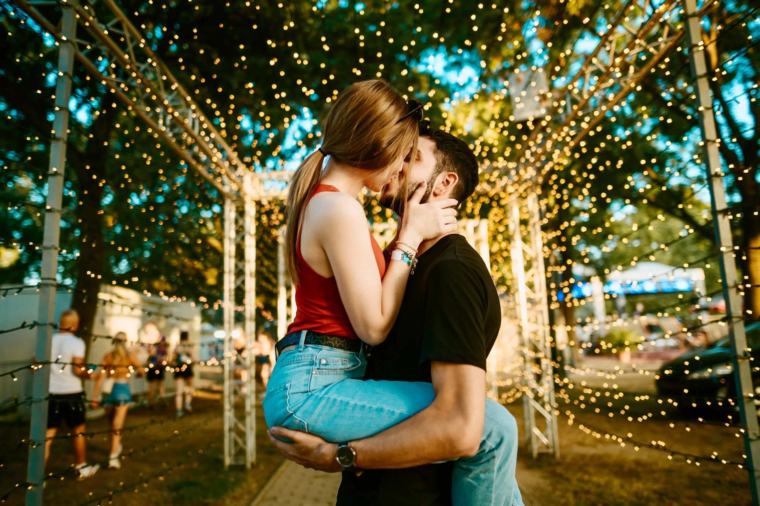 A couple kissing at an outdoor event decorated with string lights at dusk.