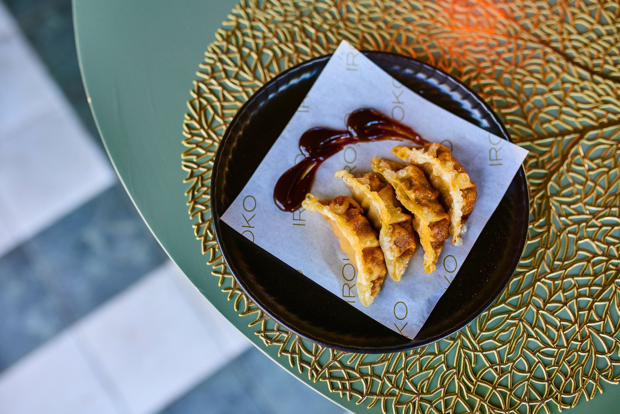 A black plate with fried dumplings drizzled with dark sauce, placed on a sheet of branded paper, resting on a green table with a decorative gold pattern.