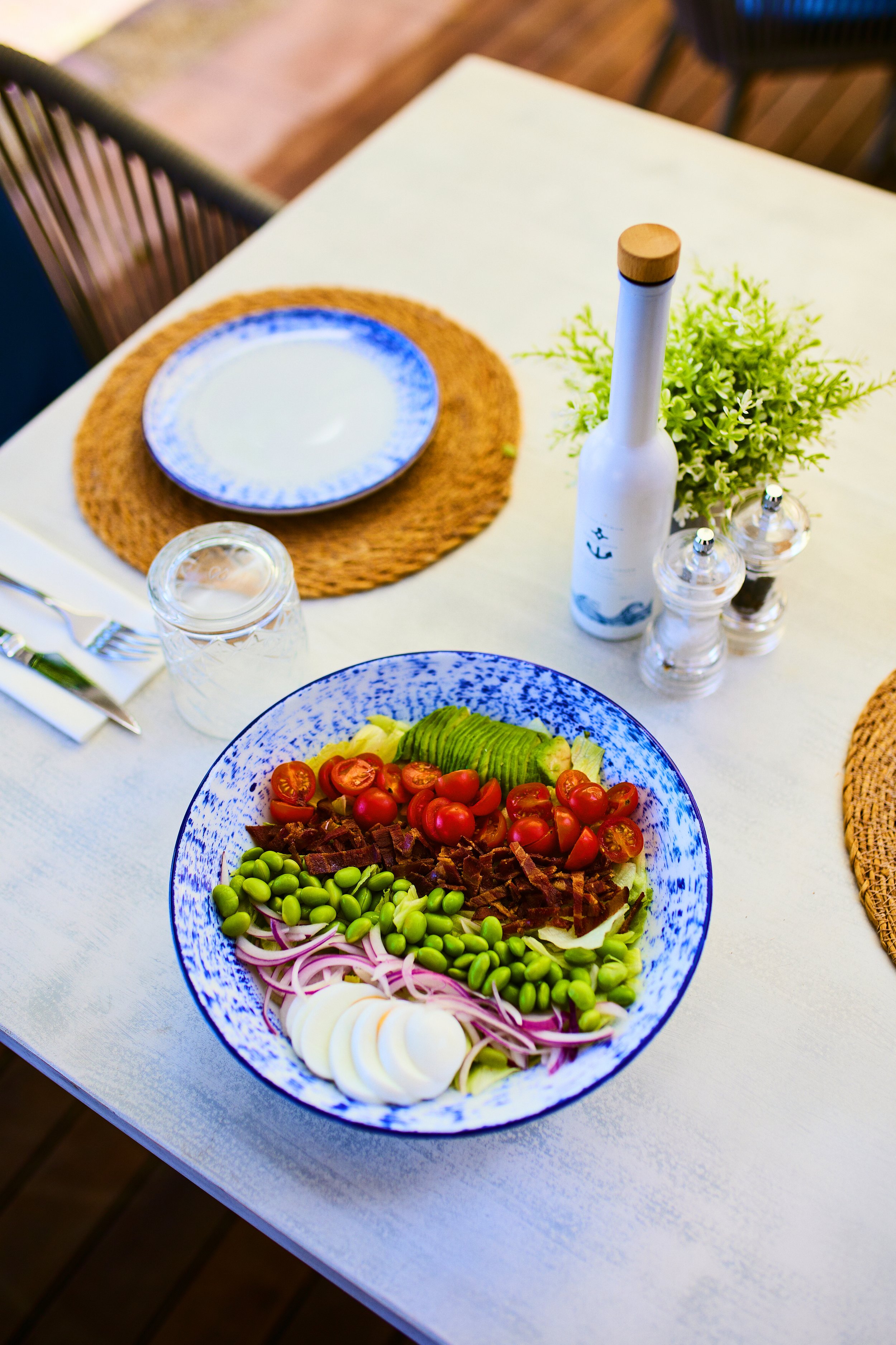 A tabletop with a salad bowl containing cherry tomatoes, avocado slices, sliced boiled eggs, onion, green beans, bacon bits, and cherry tomatoes, with wine, salt and pepper shakers, a plant, a blue and white dinner plate, a glass of water, and cutler