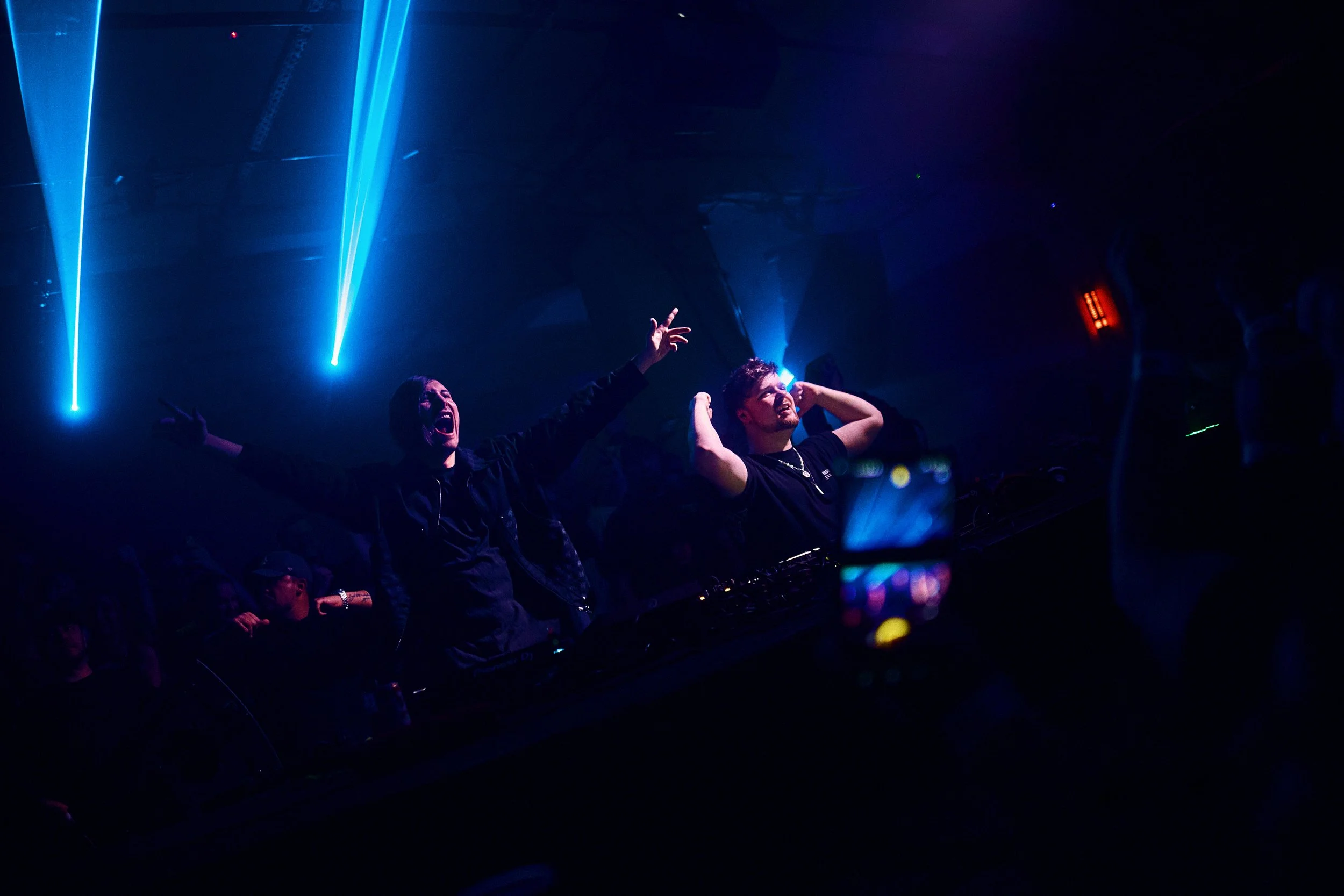 Two DJs perform at a nightclub with blue and purple lighting, one raising his arms and the other adjusting headphones, while an audience member captures the moment on a smartphone.