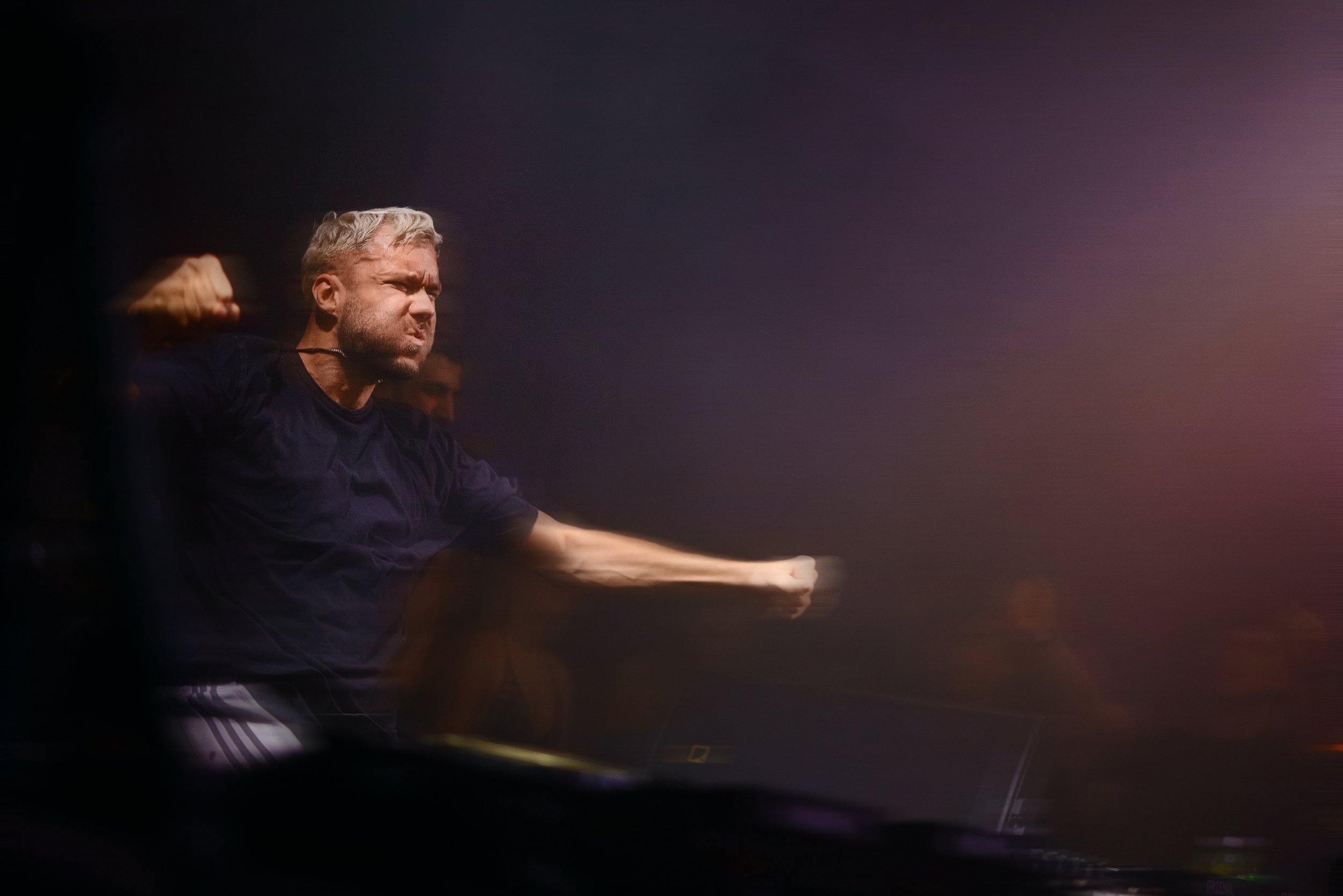 A man with blonde hair and a beard is wearing headphones and making a fist with his right arm, appearing intense and focused.