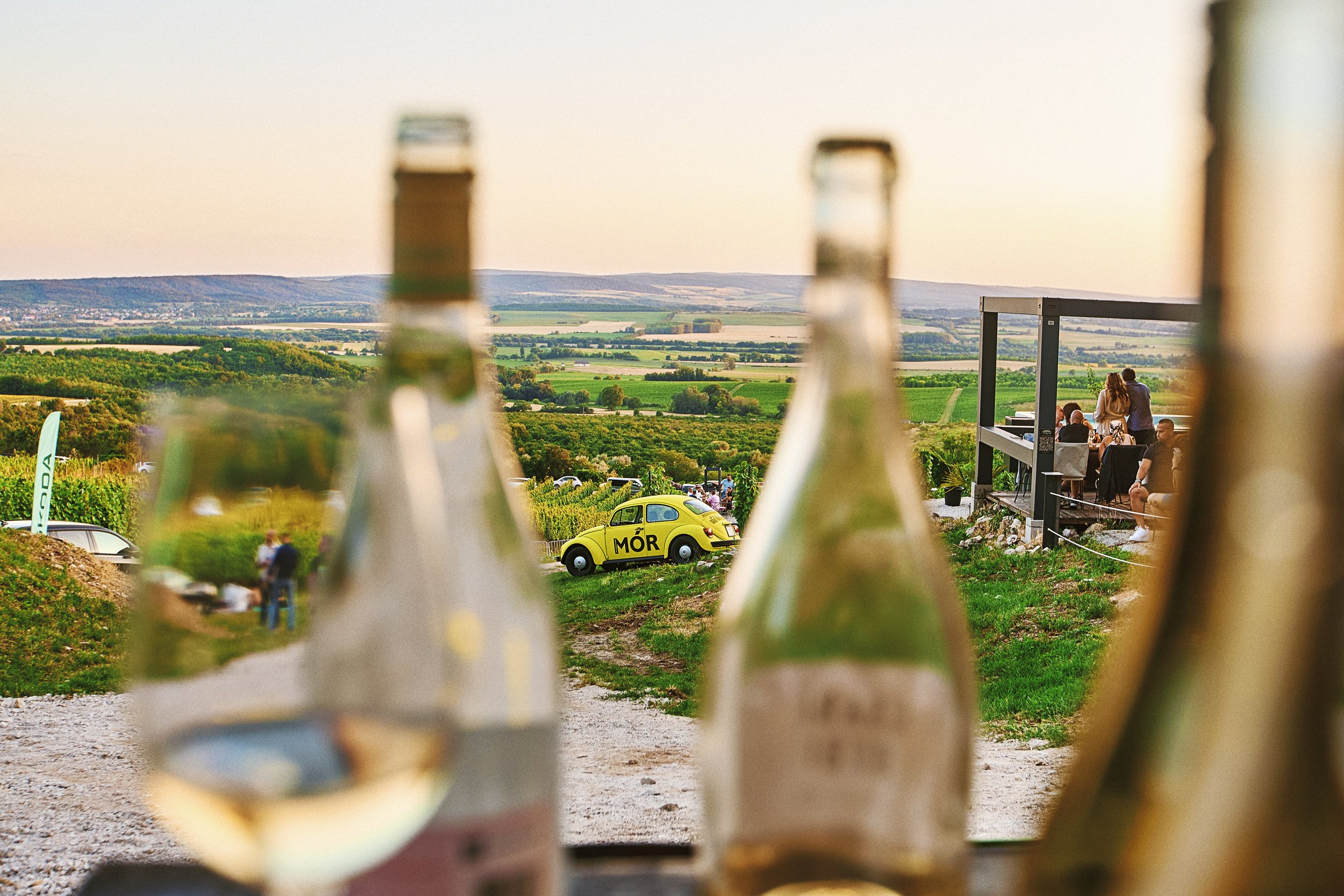 A scenic outdoor vineyard setting with a yellow vintage car and people sitting on a balcony, with wine bottles in the foreground and rolling hills in the background.