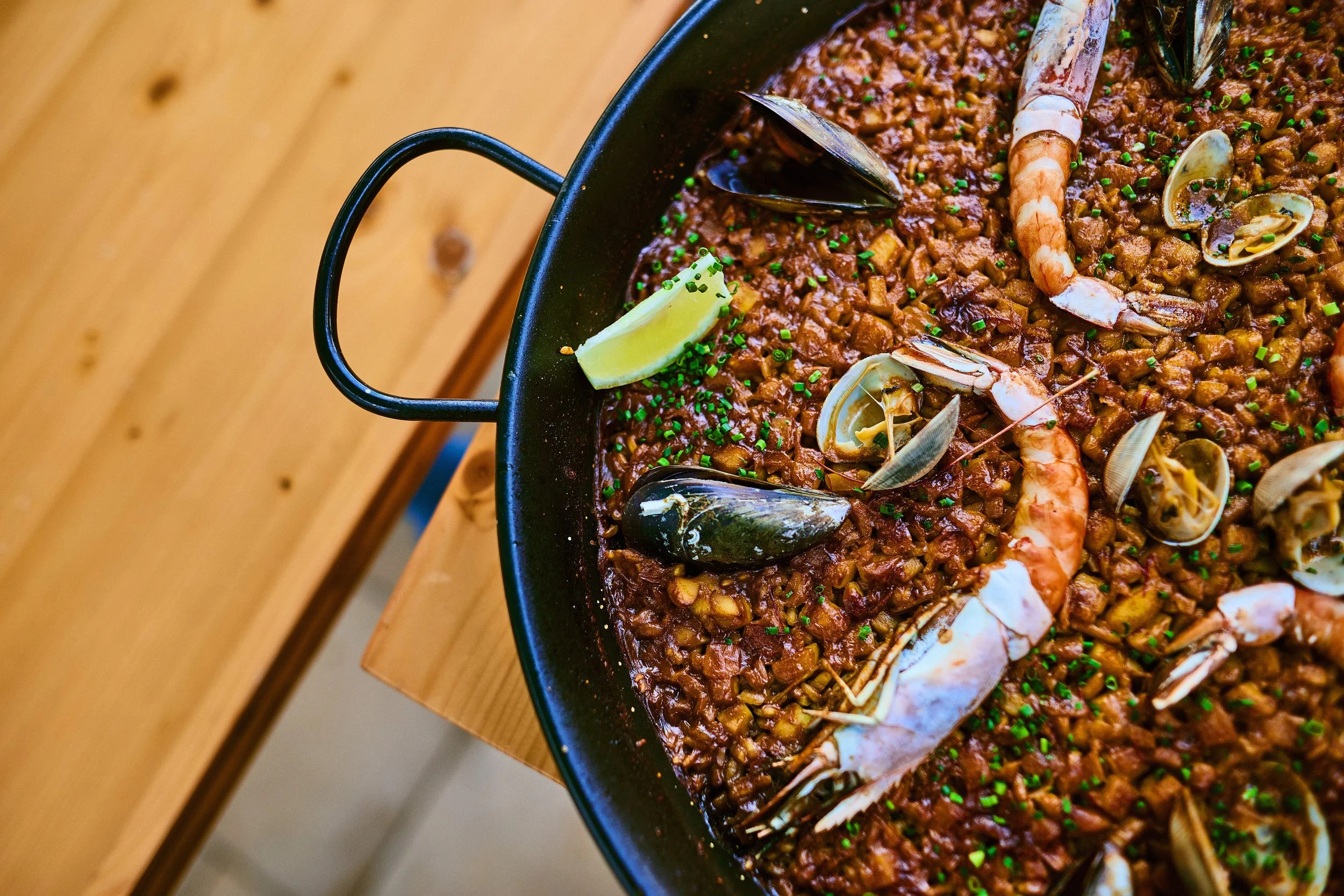 Seafood paella in a black pan with lid handle, featuring shrimp, clams, and chives, on a wooden table.
