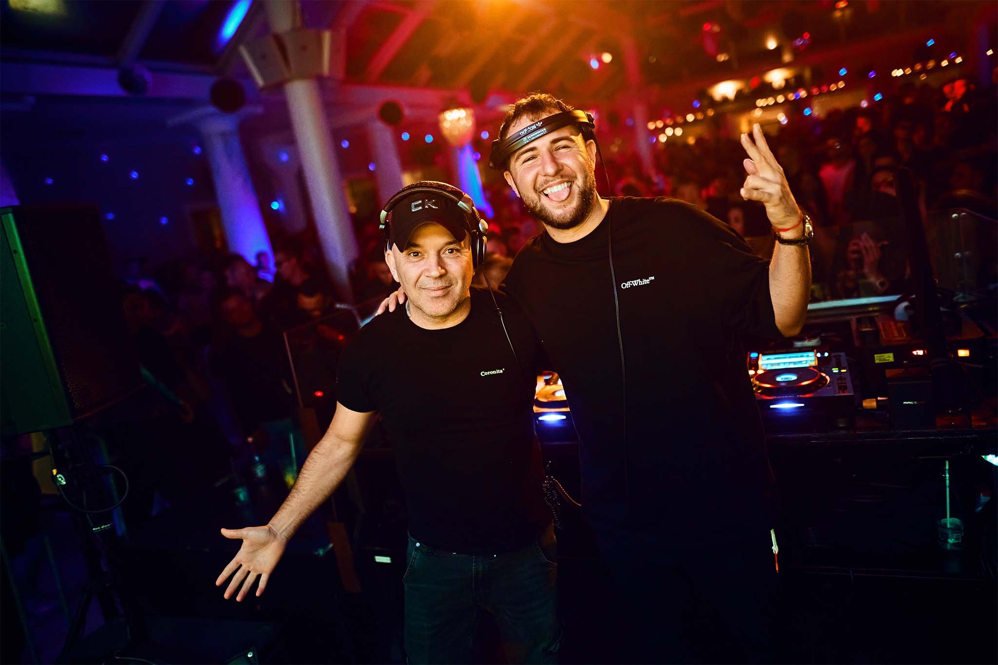 Two male DJs in a club, wearing black t-shirts and headphones, smiling and posing by the DJ equipment with a lively crowd in the background.