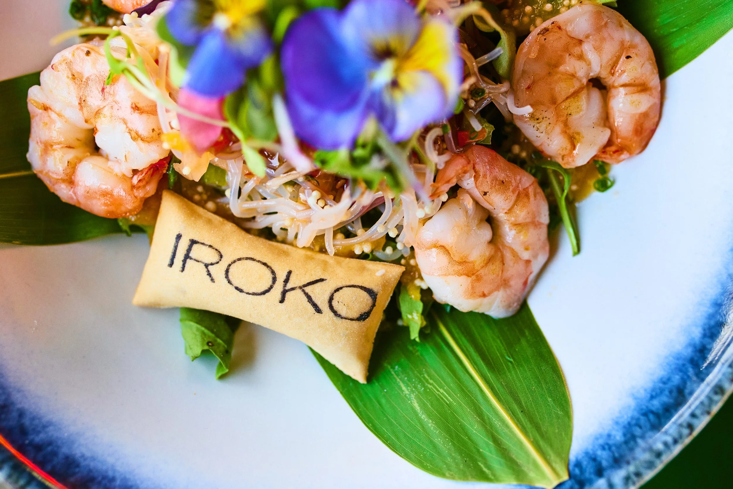 Fresh seafood dish with shrimp, noodles, purple edible flowers, and green leaves on a white plate, with a small edible sign that reads 'IROKO'.