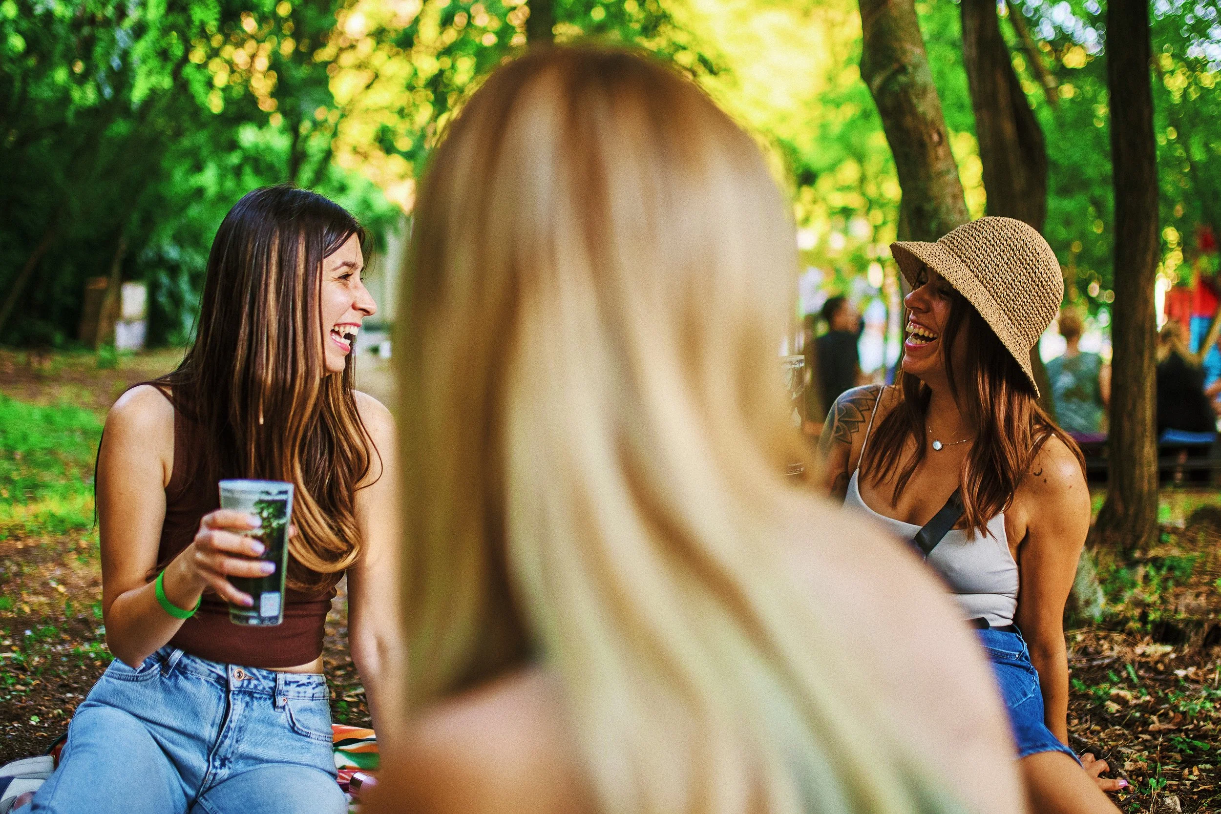 Three women laughing and talking in a park, with green trees in the background. One woman has long dark hair, another has a wide-brimmed hat, and the third has blonde hair. They appear to be enjoying a sunny day outdoors.