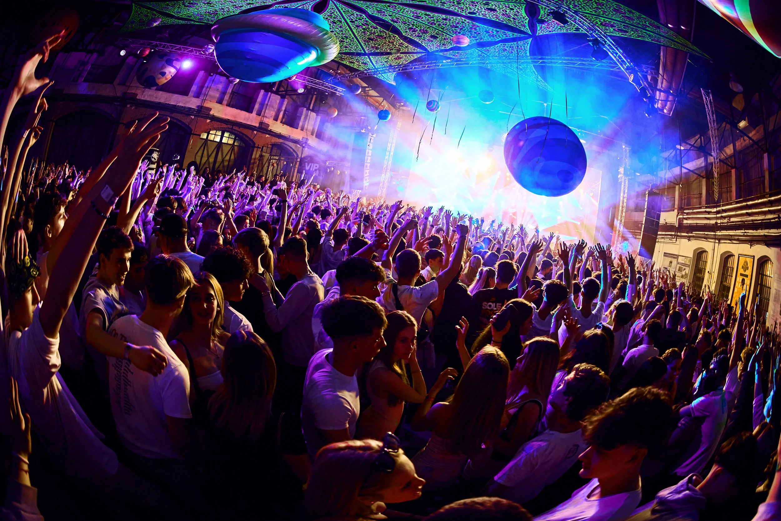 Crowd of young people at a concert or music festival with colorful lighting and large decorative objects hanging from the ceiling.