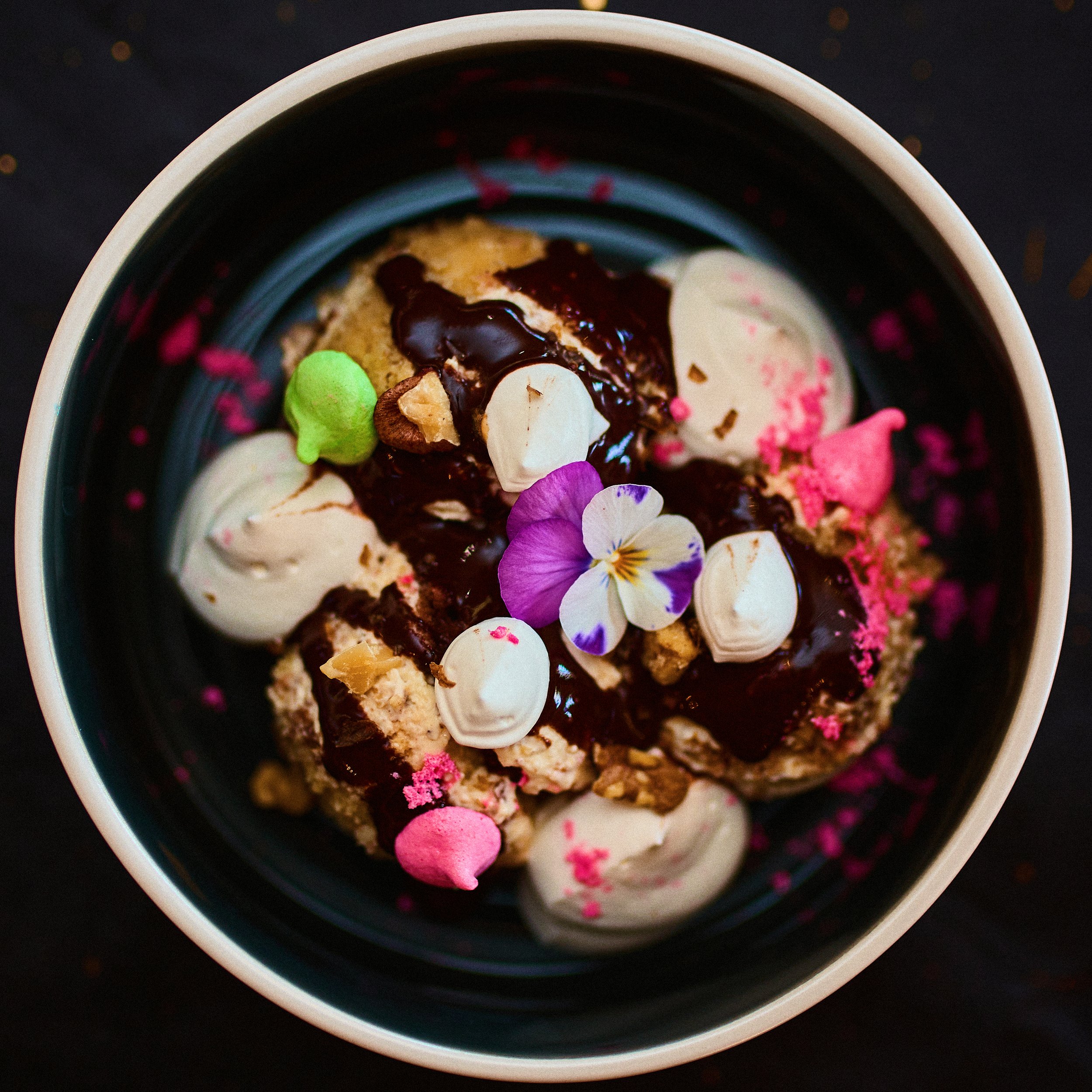 A bowl of cookies topped with chocolate drizzle, whipped cream, colorful sprinkles, edible flowers, and meringue kisses.