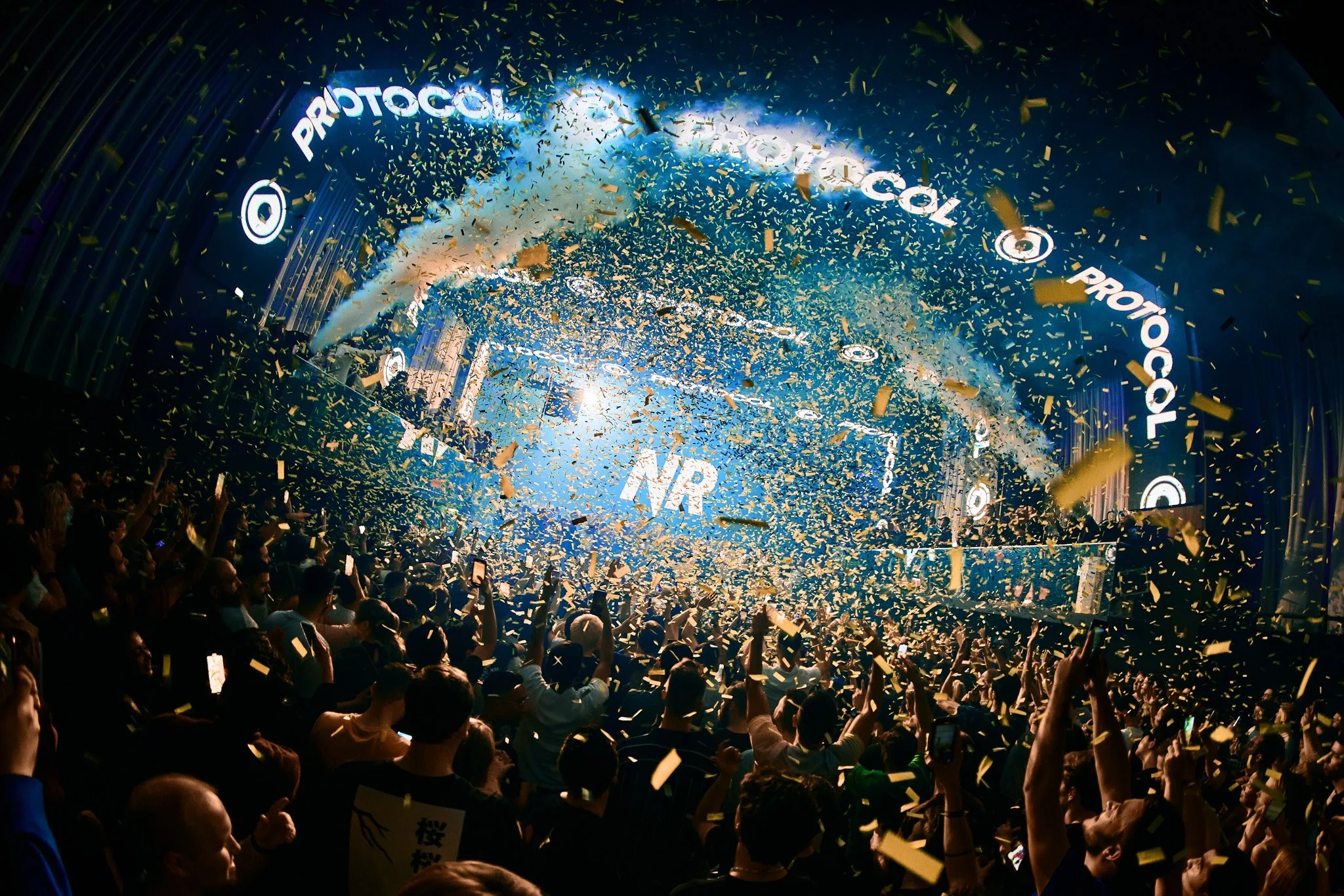 Crowd at an event with celebratory gold confetti and a large stage with the word 'PROTOCOL' displayed in lights, and a banner reading 'NR' in the background.