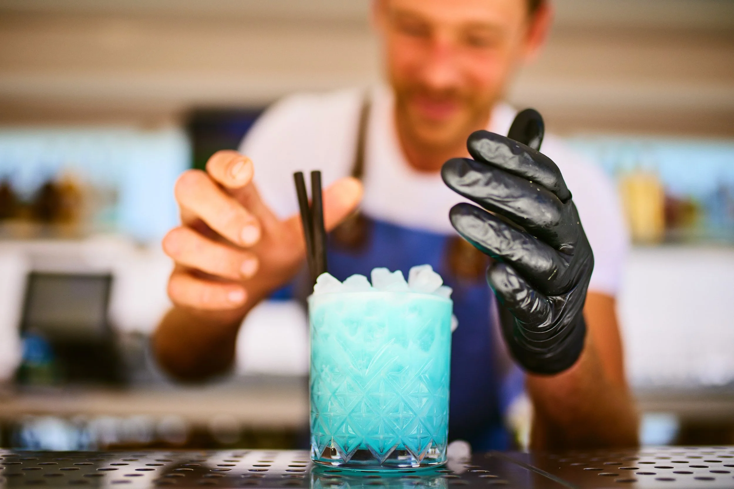 Bartender preparing a colorful blue cocktail with ice, garnished with two black straws, in a patterned glass, wearing black gloves, and smiling at a bar.