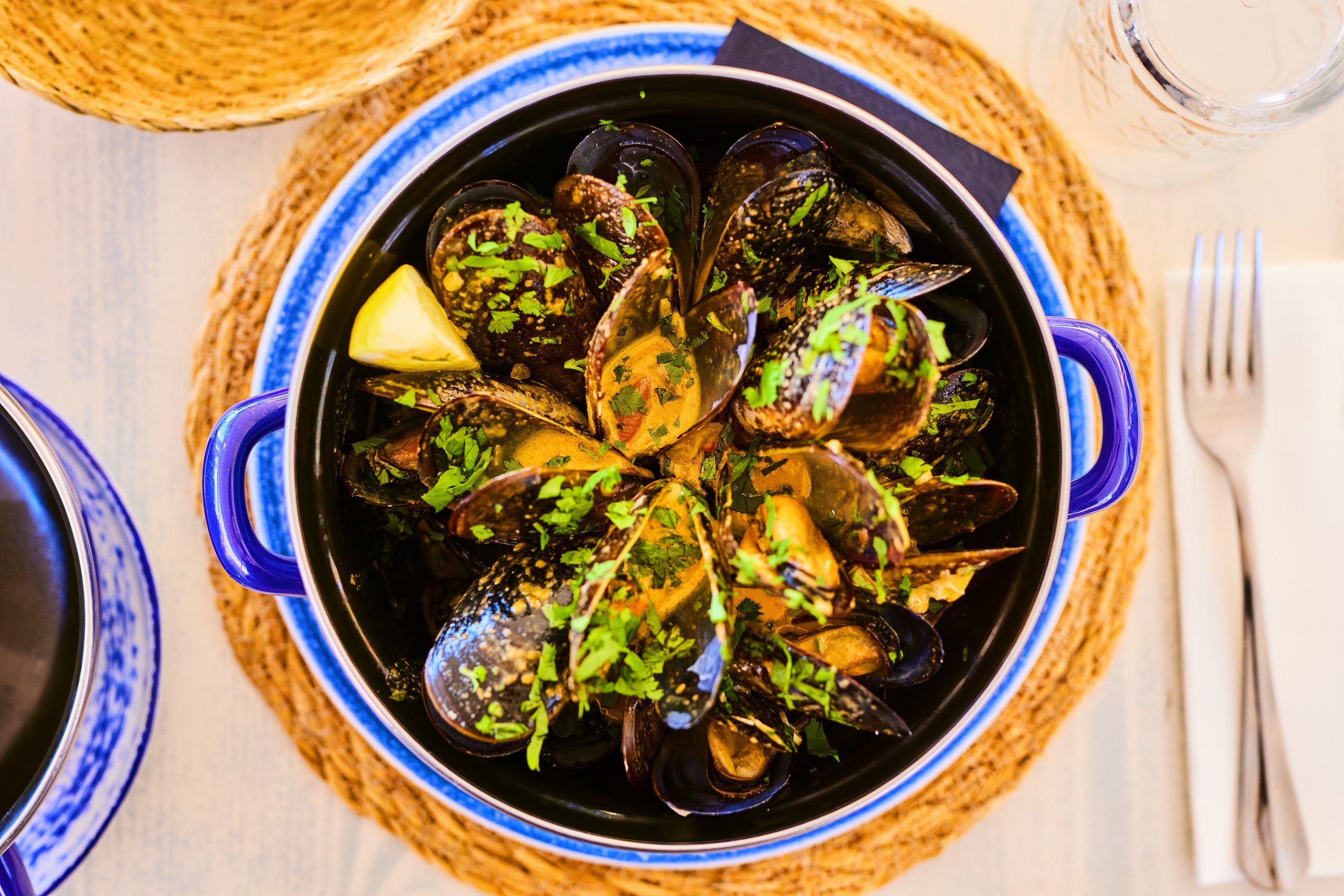 A bowl of cooked mussels garnished with chopped parsley, served with lemon wedges. The bowl is placed on a woven placemat on a dining table.
