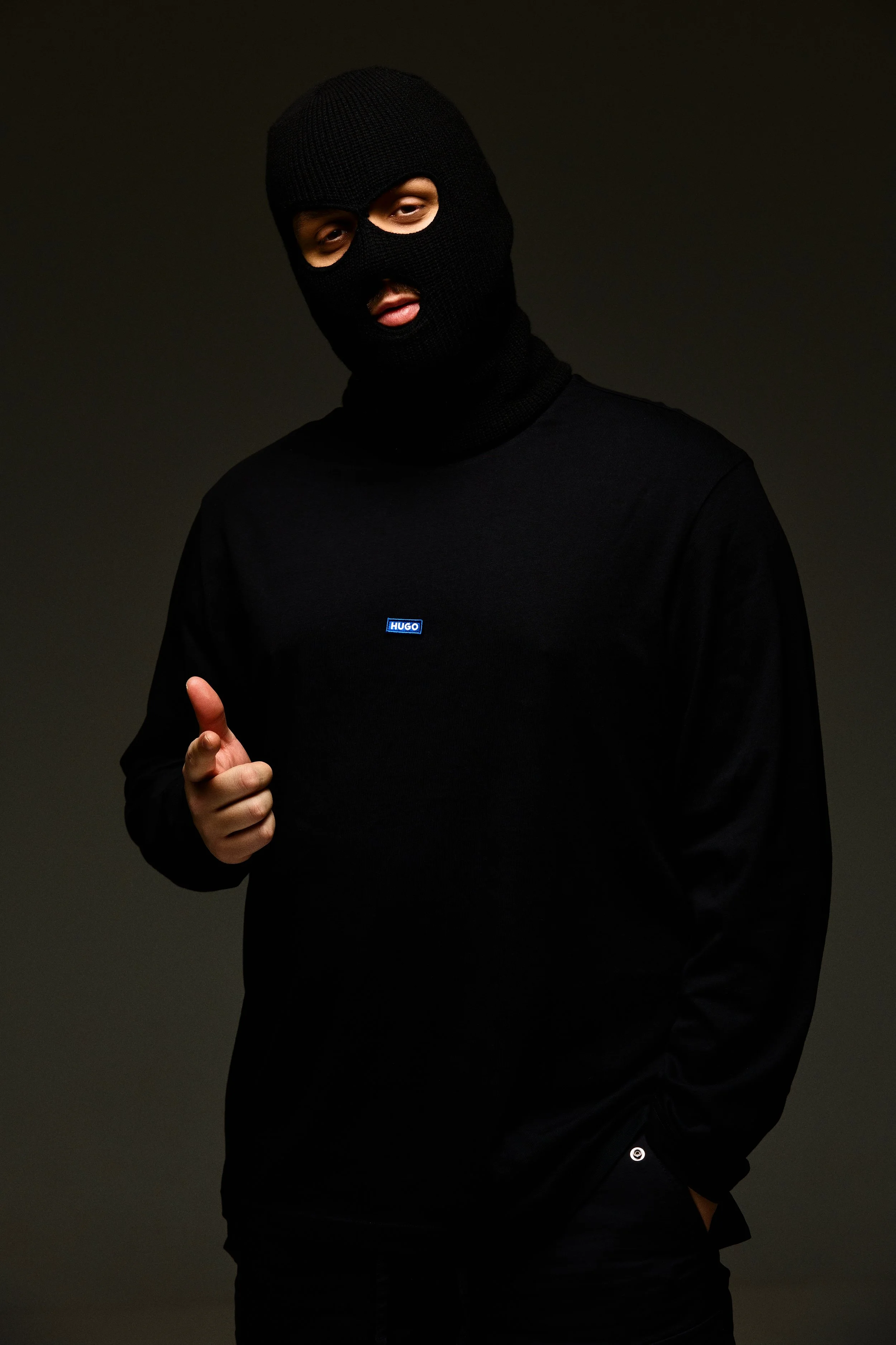 A person wearing a black ski mask, black long-sleeve shirt with a small blue logo, and black pants, pointing with their right hand and standing against a dark background.