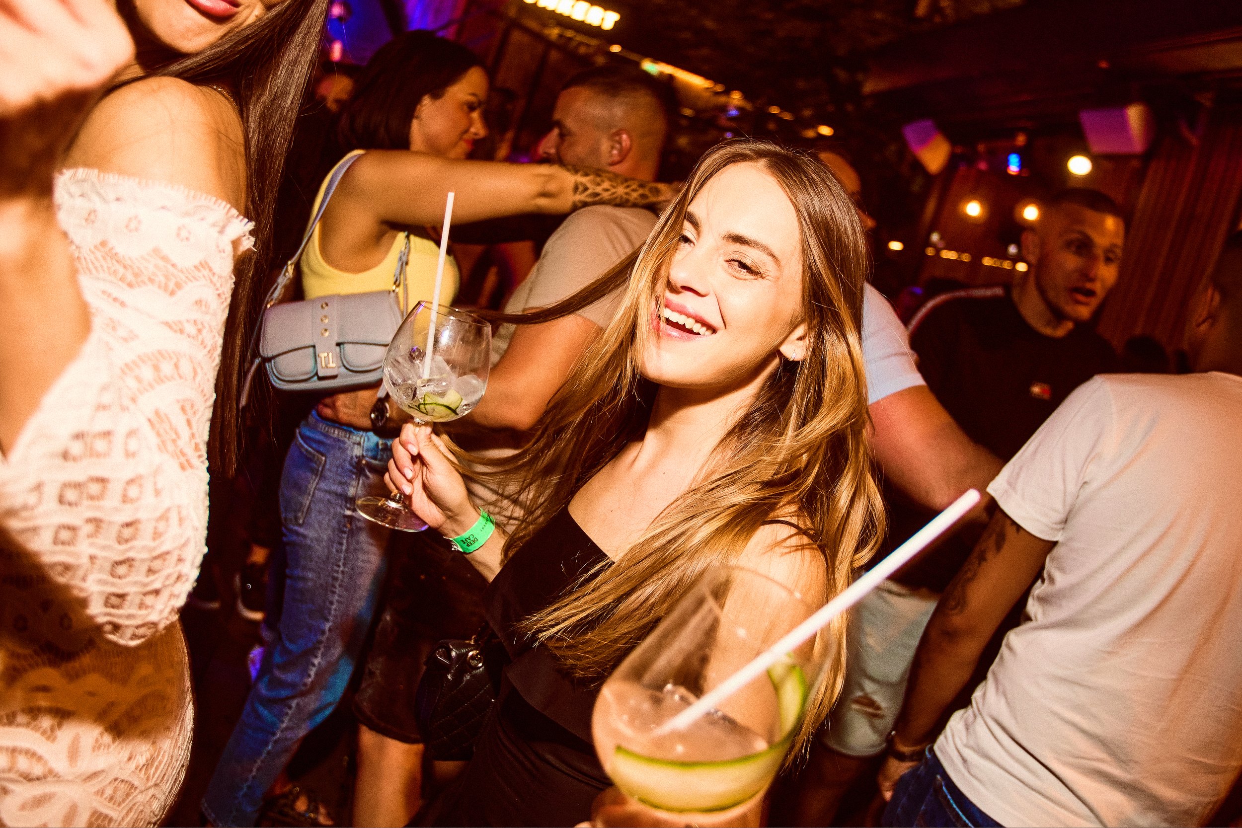 Young woman with long blonde hair smiling and winking while holding a cocktail at a lively party.