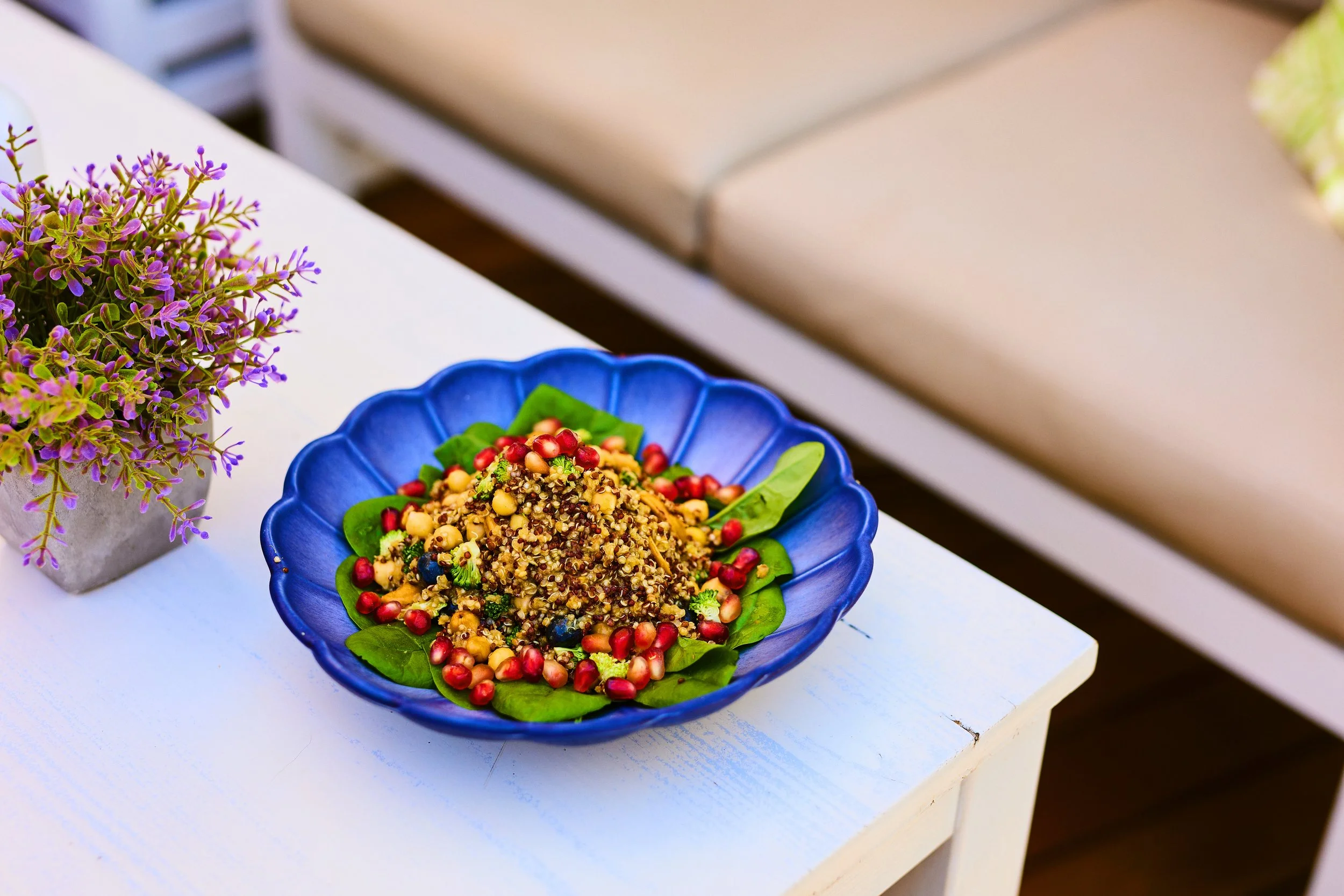 Colorful quinoa salad with pomegranate seeds and leafy greens in a blue bowl on a white table next to a pot of purple flowers.