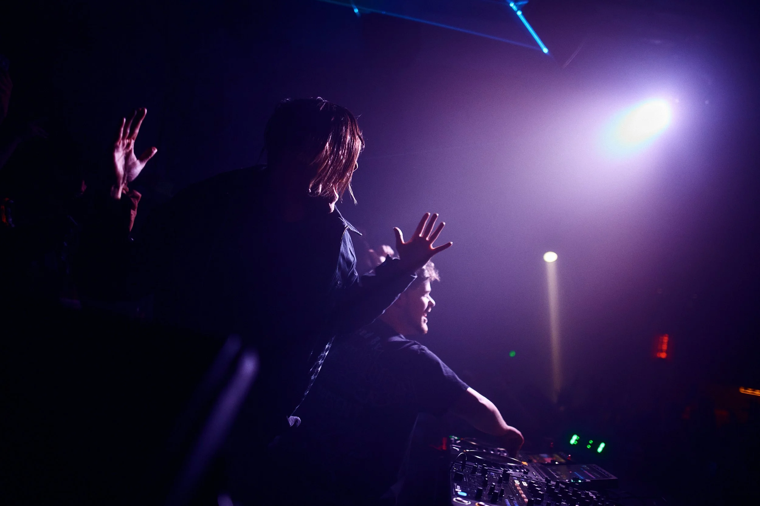 Two DJs performing at a concert or nightclub, with one DJ operating a mixer and the other with hands raised, under colorful stage lighting.