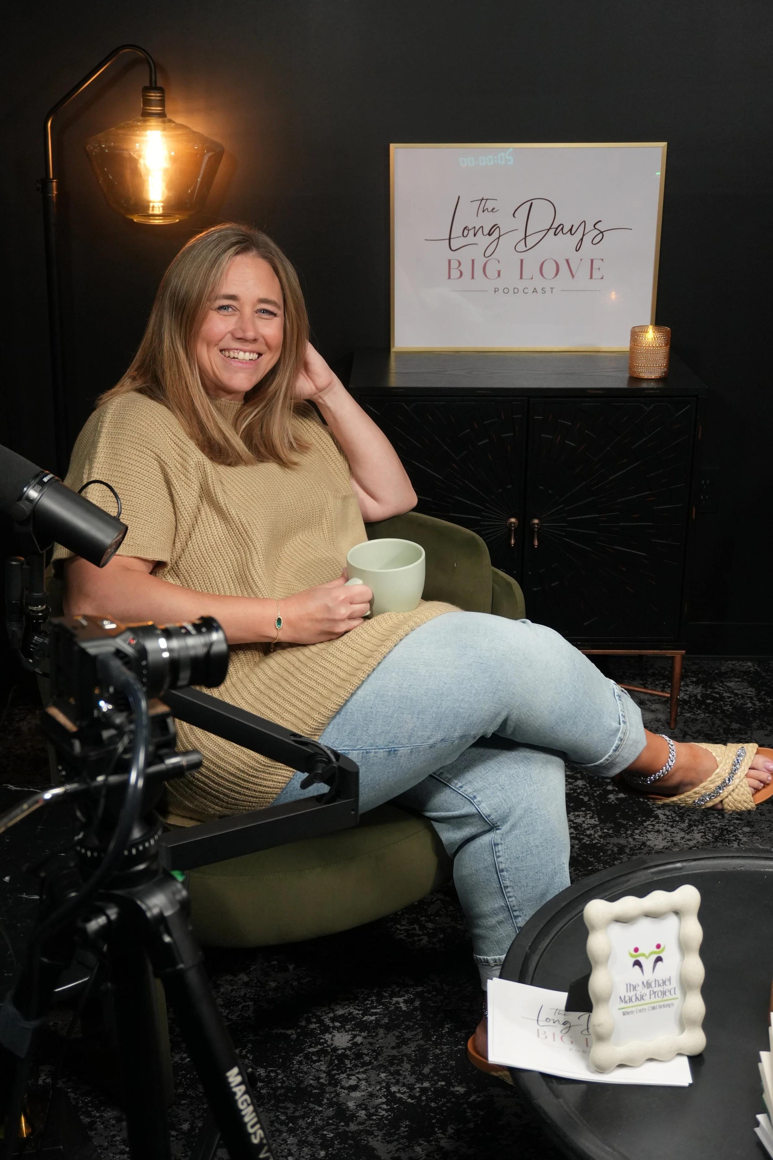 Woman sitting on a green chair, smiling, holding a white mug, in a recording studio with a black wall, a sign that reads 'The Long Days Big Love Podcast', and a lit candle on a black cabinet.