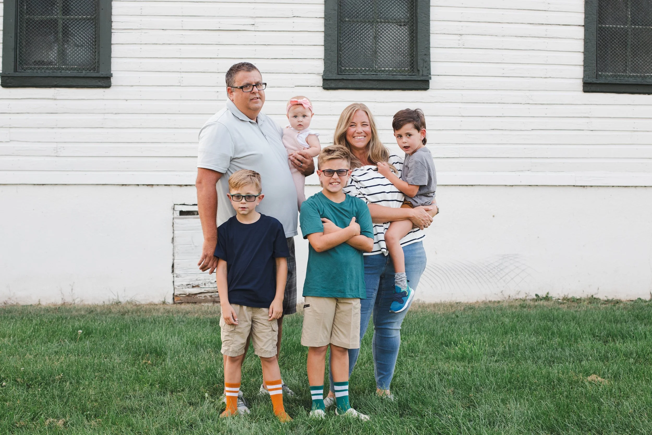 A family of six standing outside in front of a white wooden building with black window frames. The family includes a man, woman, four children, and a baby girl. The children and adult are wearing casual clothing and colorful socks, and they are posed on a grassy lawn.