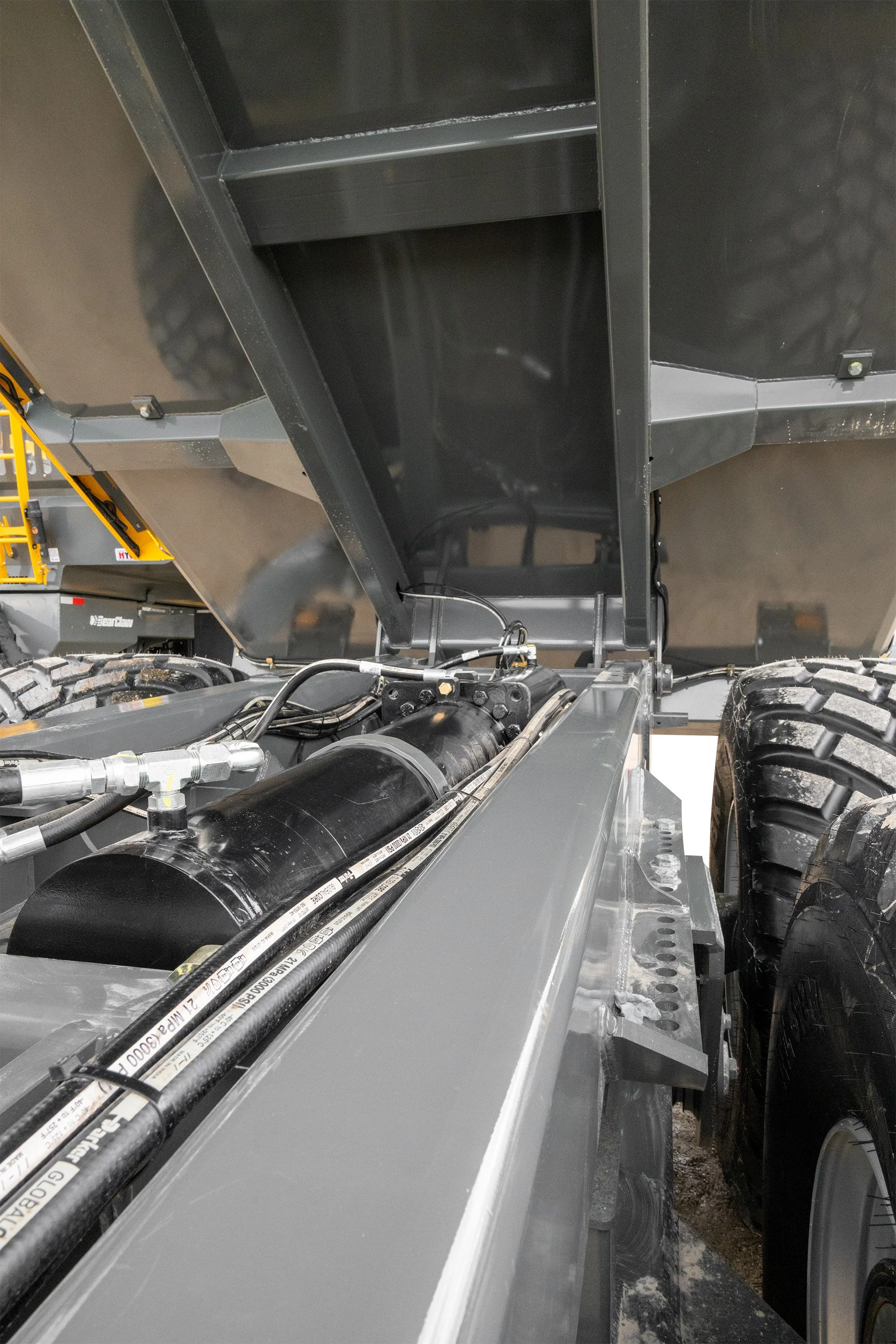 Close-up view of a large industrial dump truck focusing on the underside and hydraulic systems, with black hydraulic hoses and parts visible, as well as a portion of the truck's tire.