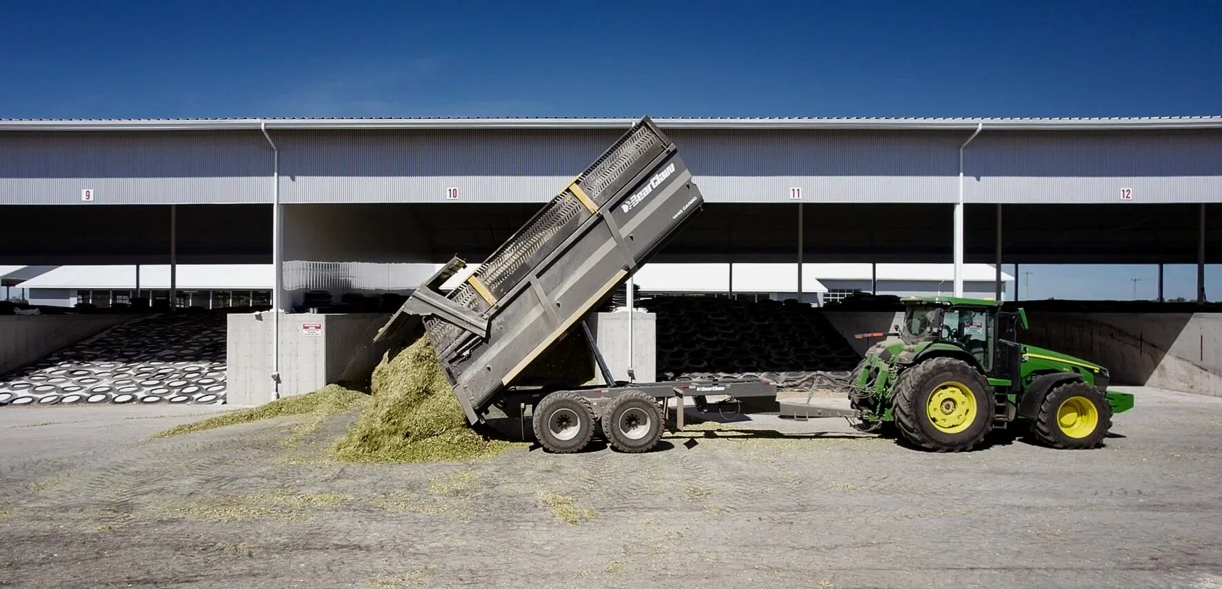 A green tractor dumping feed into a storage area at a farm with a large covered structure overhead.