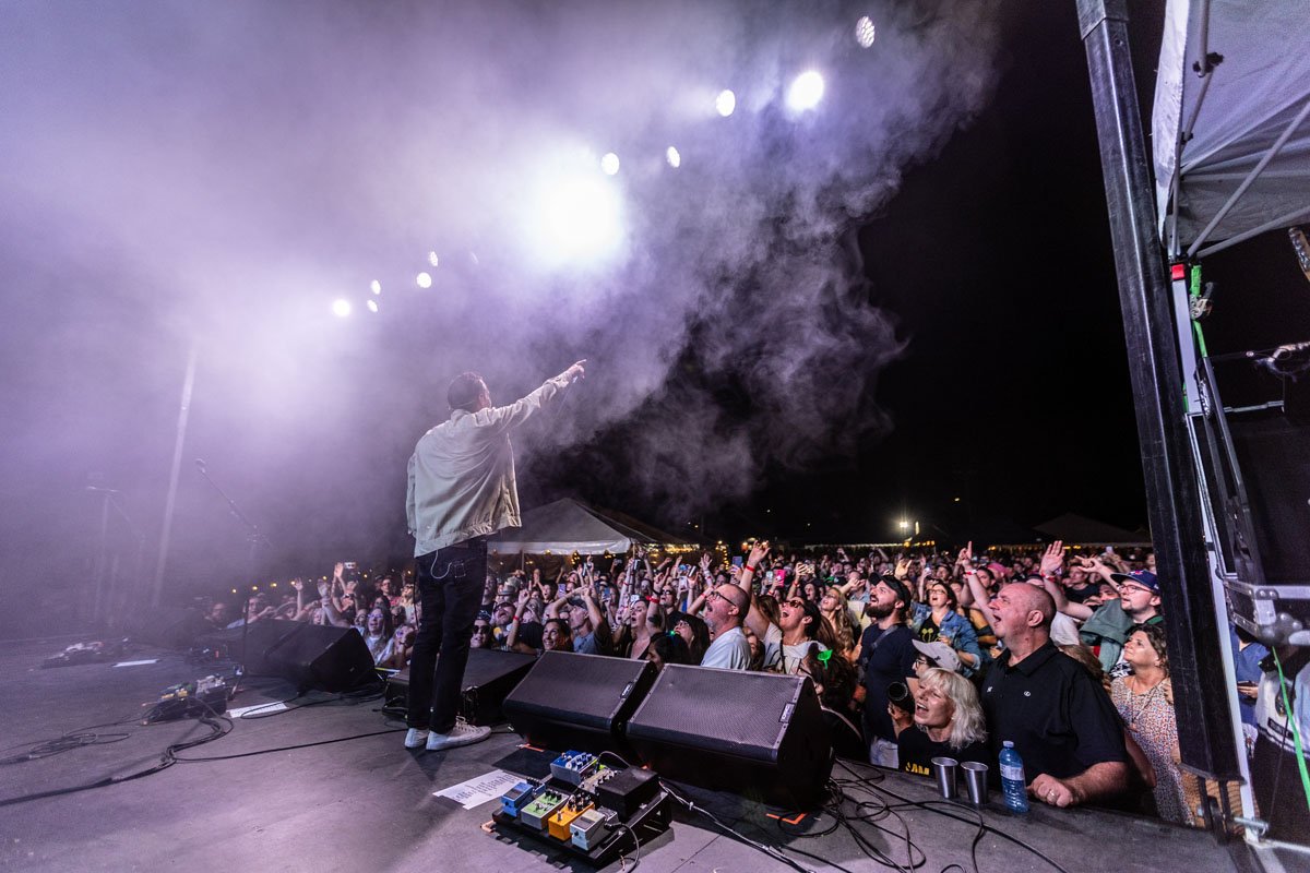 Performer on stage pointing to crowd at night concert with smoke and stage lights, audience raising hands and enjoying music.