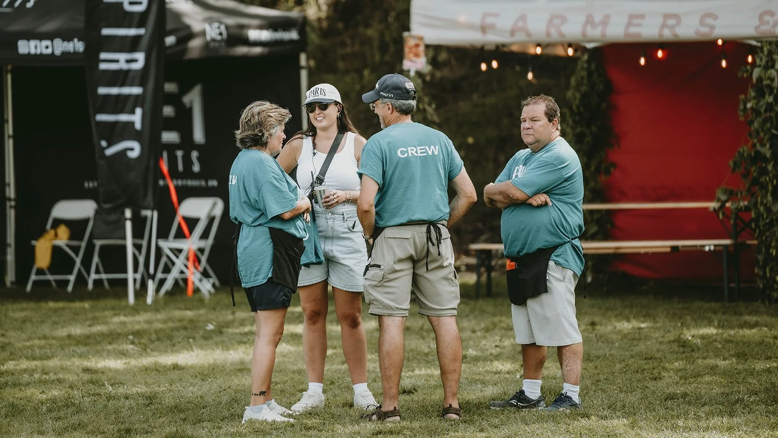 Four people standing on grass in front of tents, engaged in conversation; three wearing blue shirts, one woman in white tank top and shorts, two men with "CREW" shirts.