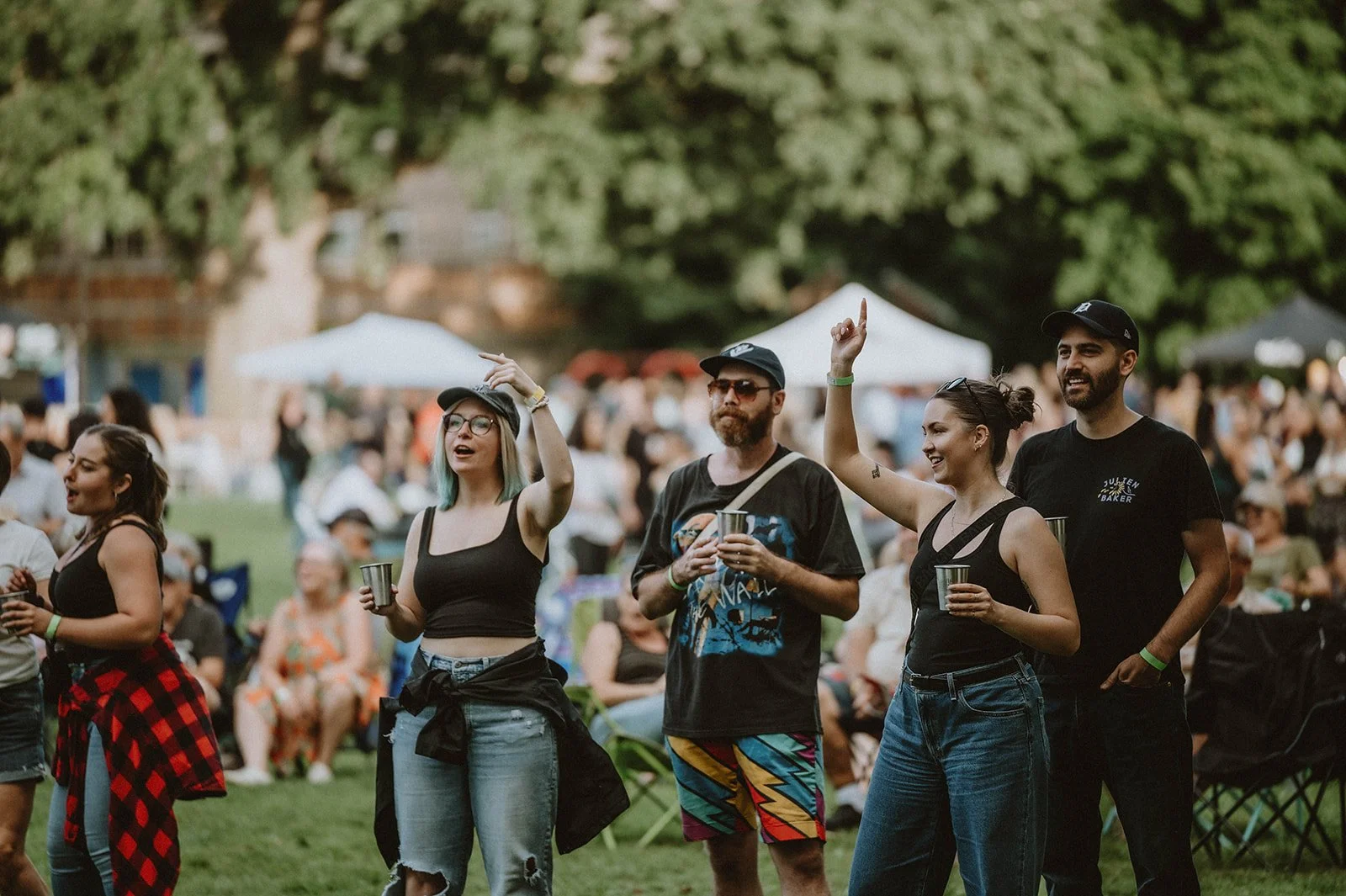 Group of five people standing outdoors at a festival, some raising their hands and holding drinks, with a crowd and trees in the background.