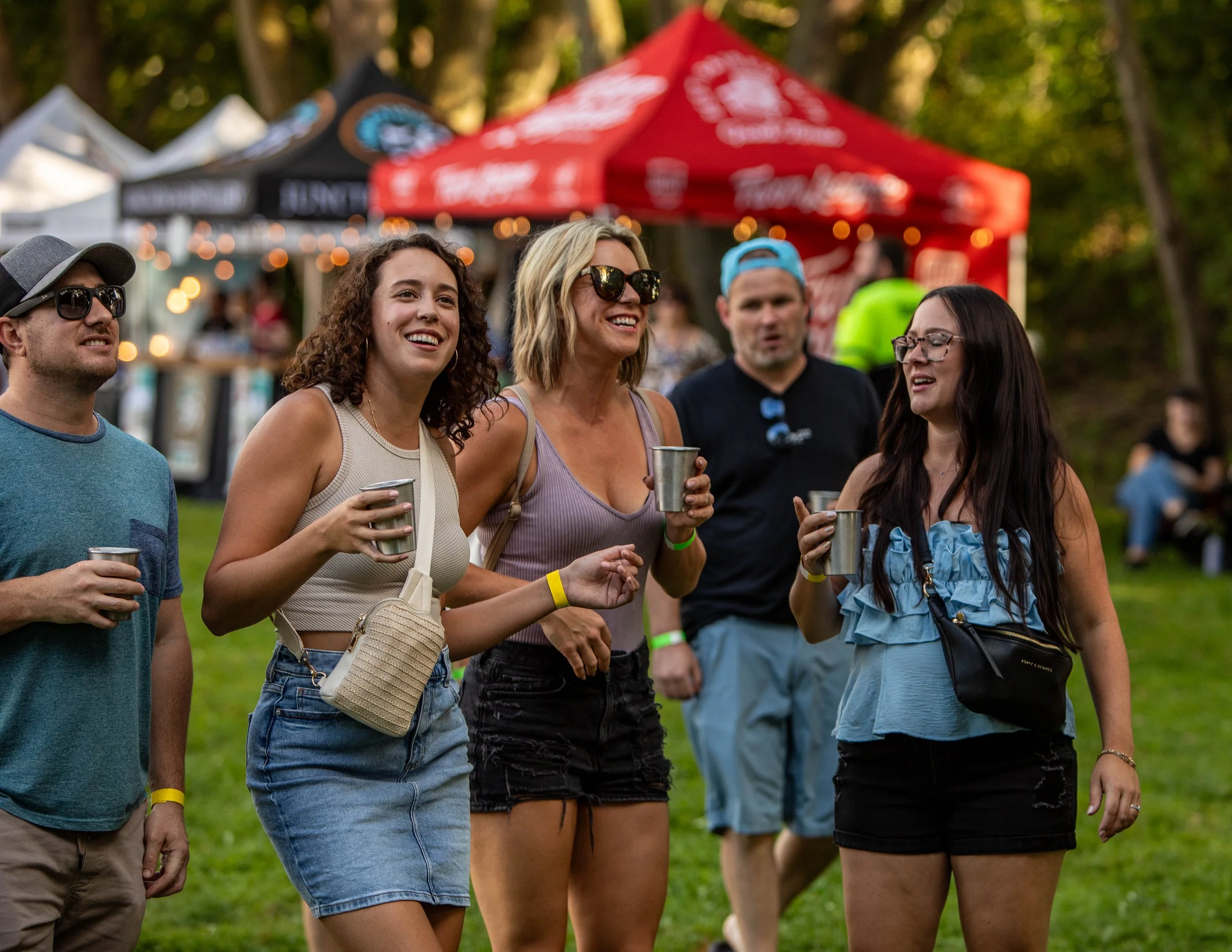 A group of five people, three women and two men, enjoying drinks and socializing at an outdoor event with tents and string lights in the background.