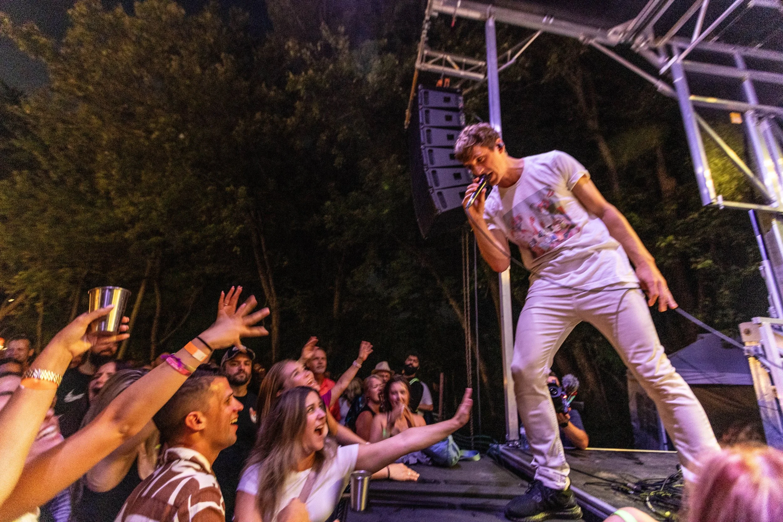 A performer singing on stage at night while an audience reaches out towards them at an outdoor concert.