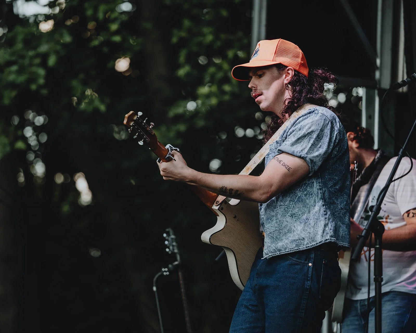A musician playing an acoustic guitar on stage outdoors, wearing a denim shirt, orange cap, and having long curly hair, with trees in the background.