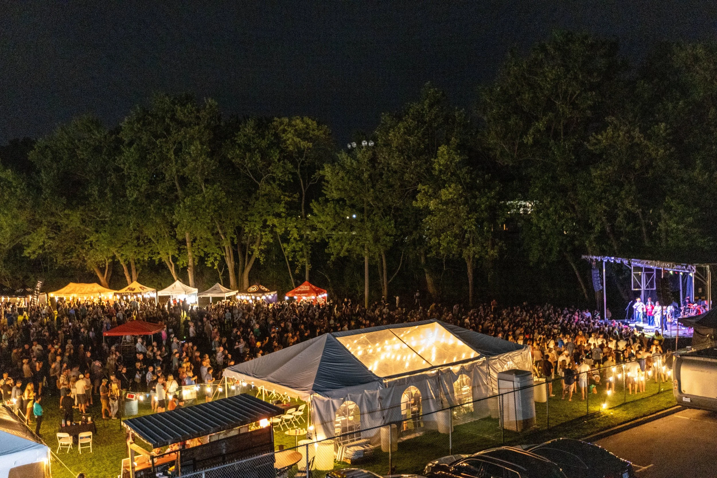 Nighttime outdoor concert with a large crowd gathered in front of a stage with musicians, surrounded by trees and illuminated tents.