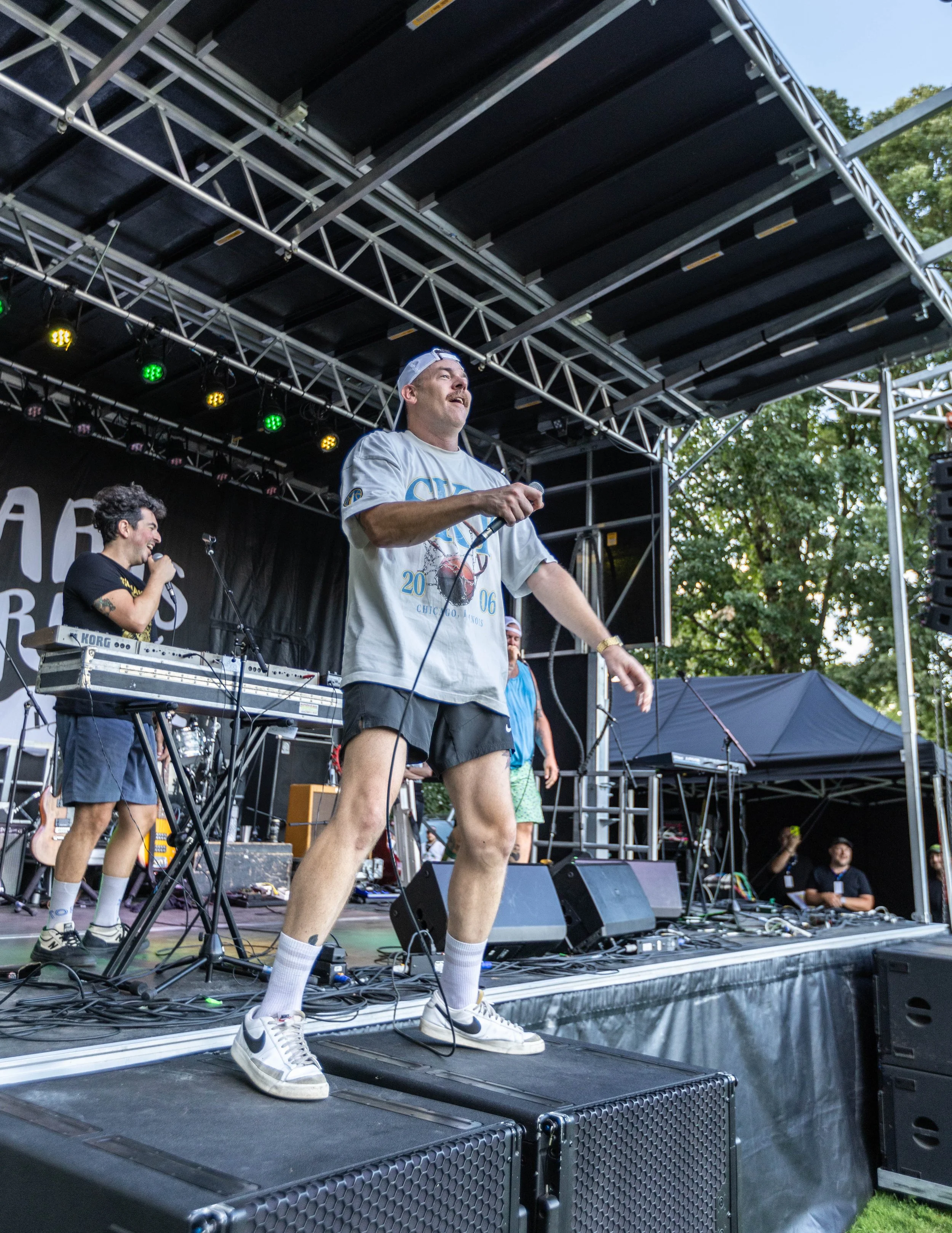 Performers on stage at an outdoor concert, with one man wearing shorts, a t-shirt, sneakers, and cap, holding a microphone.
