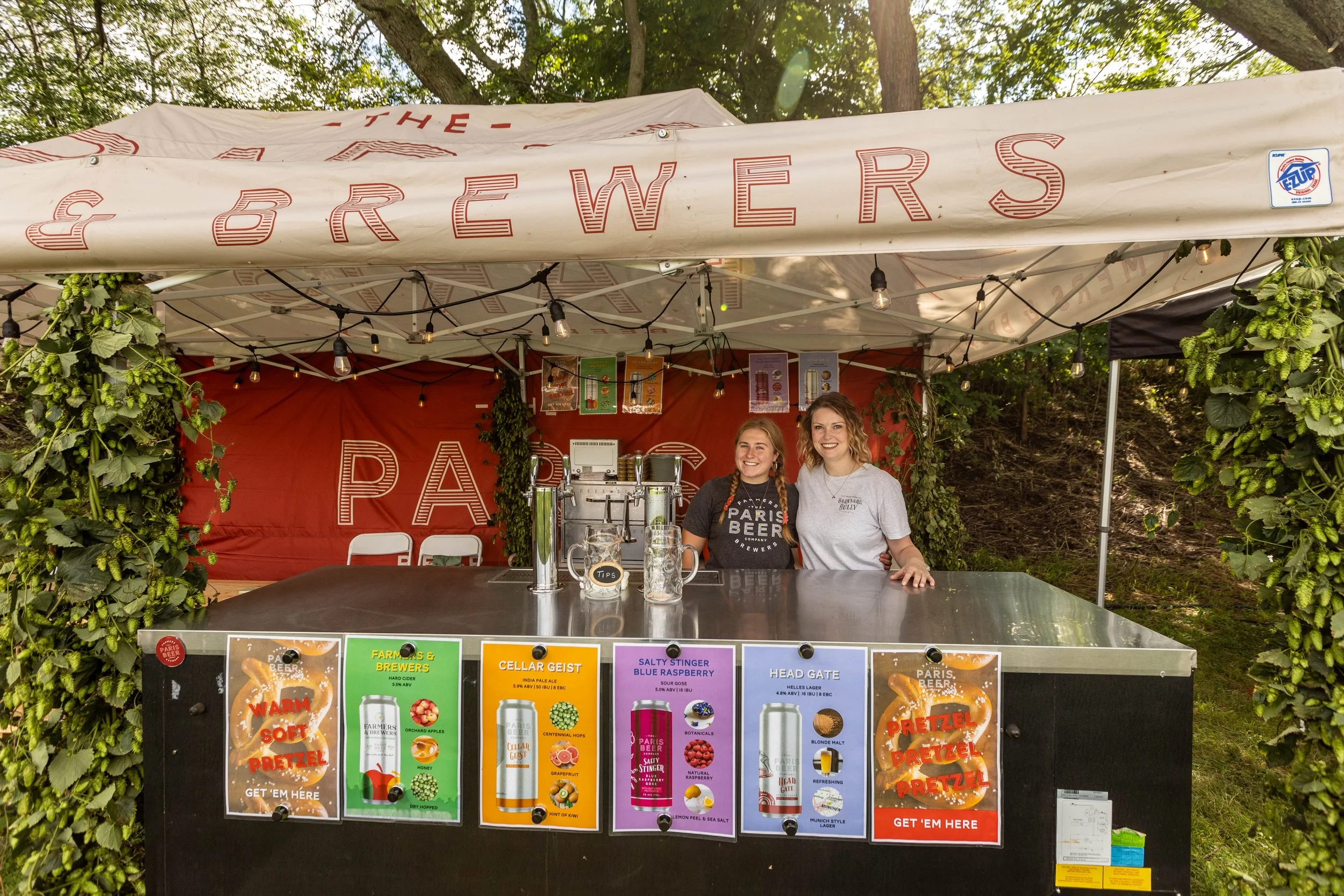 Two women standing behind a beer stand under a white canopy with the word 'BREWERS' on it. The stand features colorful posters advertising various beer flavors and pretzels, with one smiling woman wearing a black 'Paris Beer' T-shirt and another in a