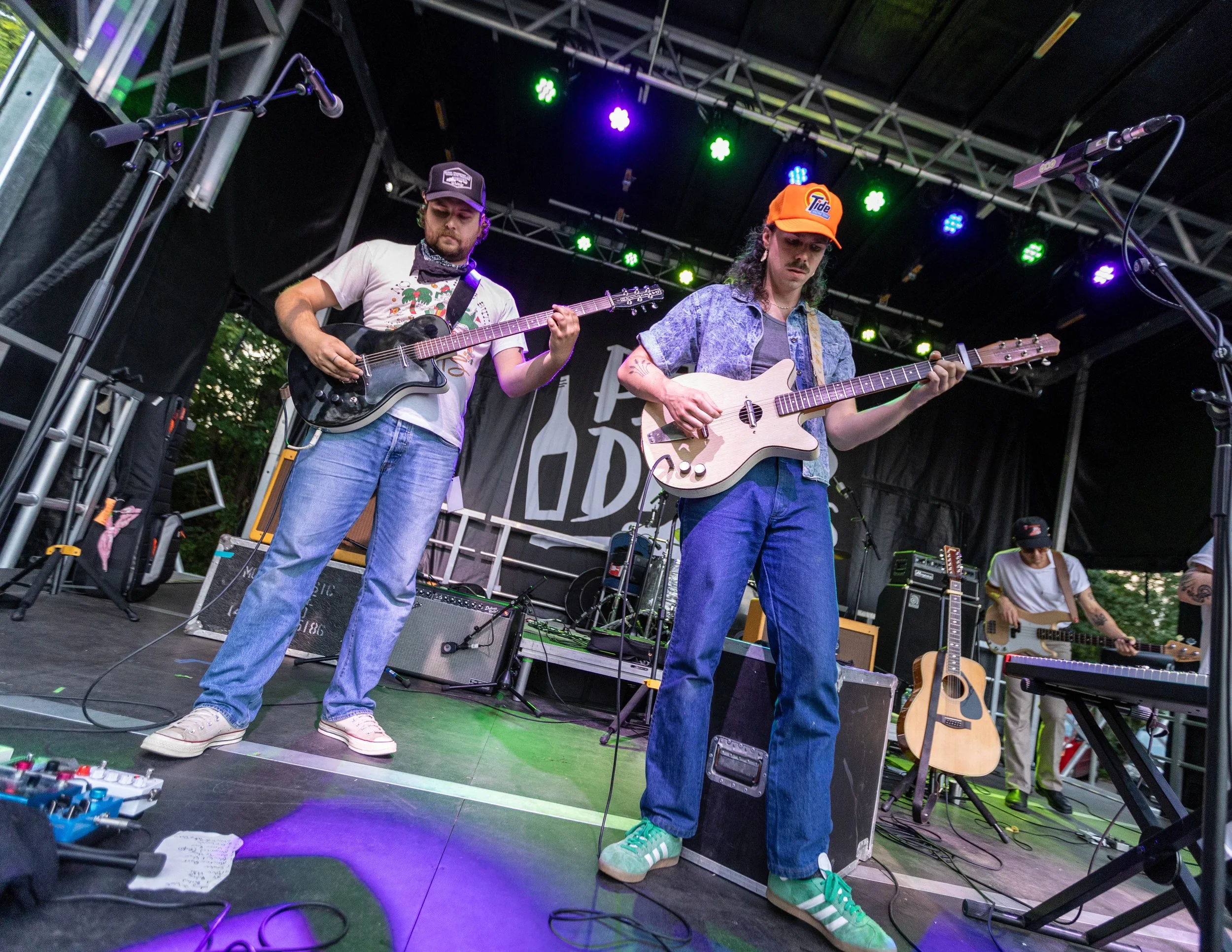 Two musicians playing electric guitars on stage during a live outdoor concert, with colorful stage lights and additional band members in the background.