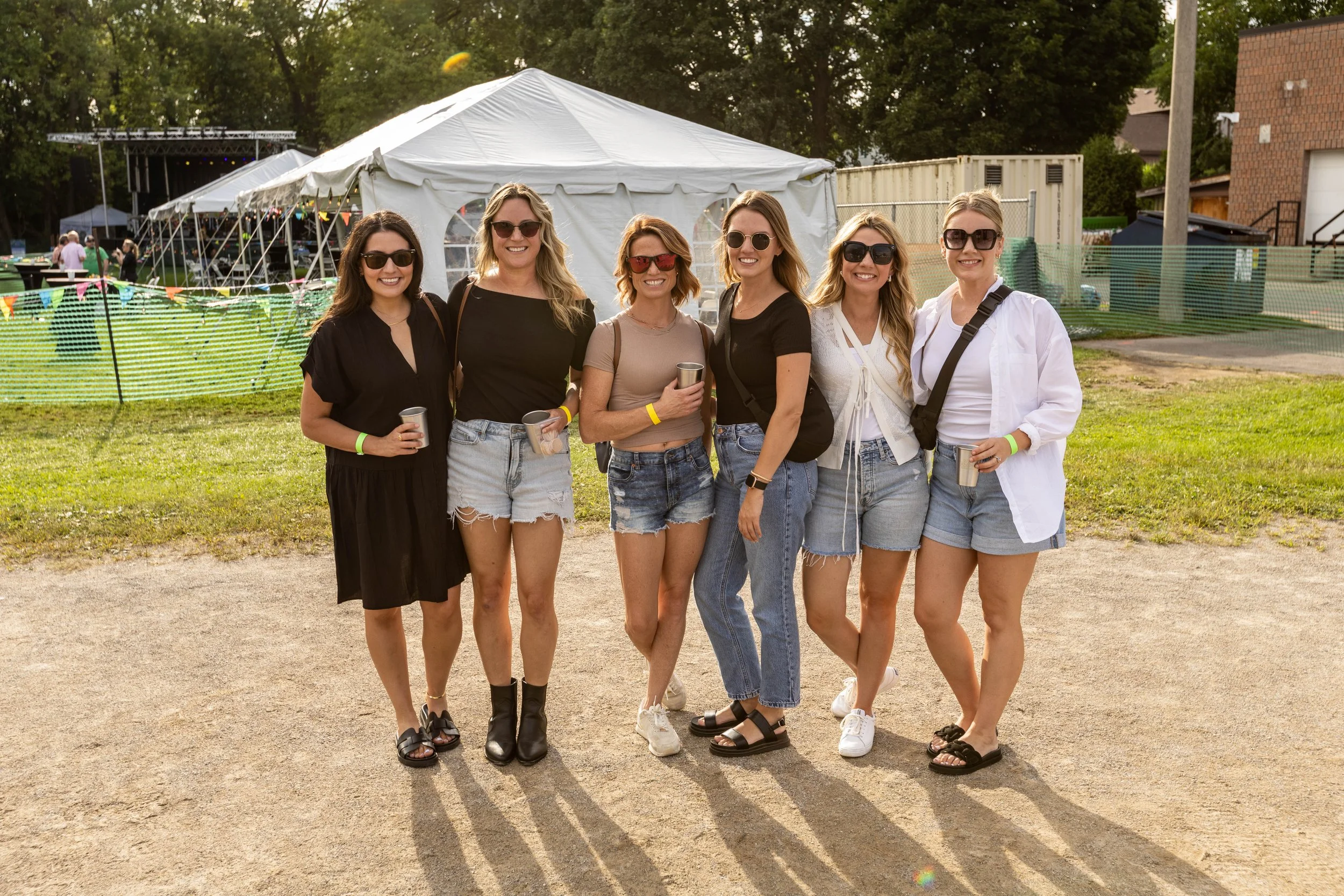 Six women standing together outdoors at a festival or gathering, smiling, with a large tent, people, and trees in the background.