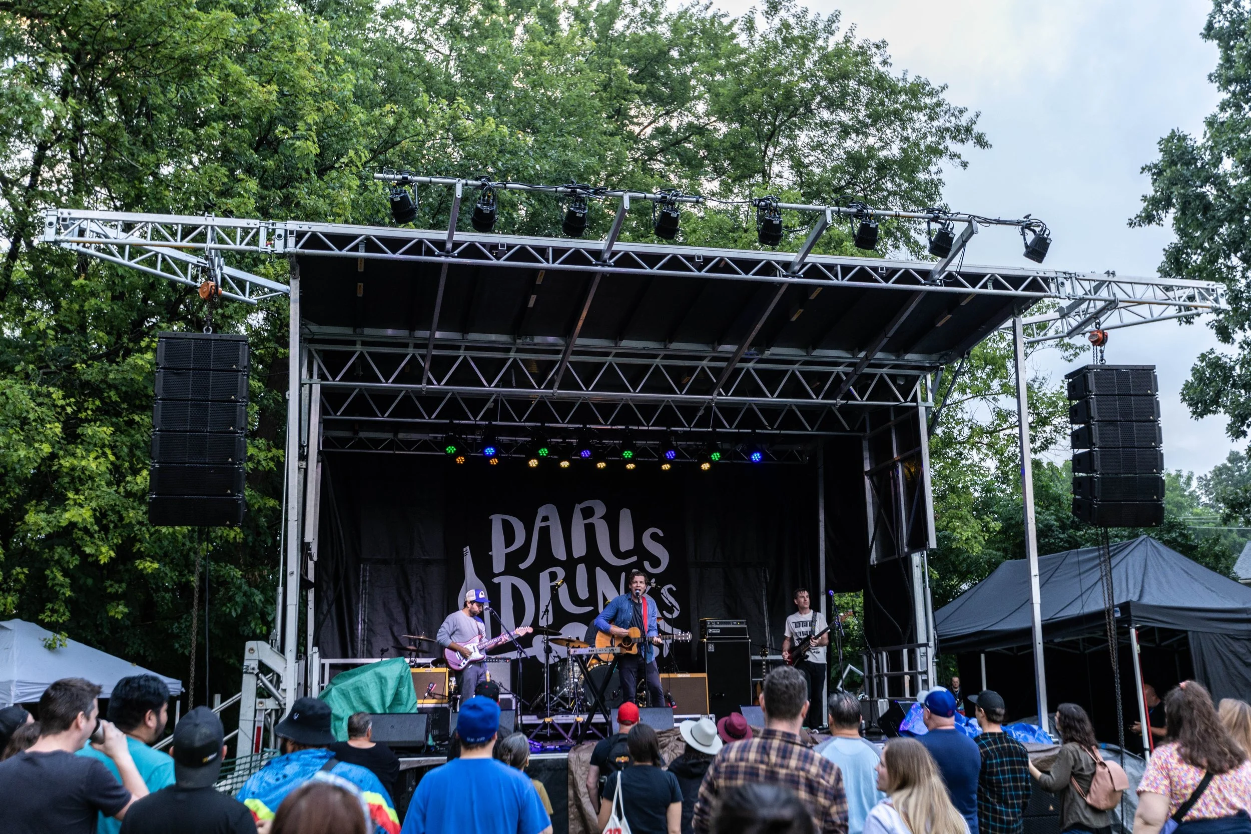 Outdoor concert stage with a band performing and an audience watching, surrounded by trees.
